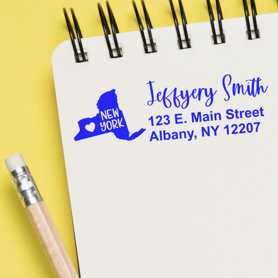 New York State Love Personalized Address Stamp on a notepad, featuring a blue state outline with a heart, personalized with name and address. Pencil nearby on a yellow background.