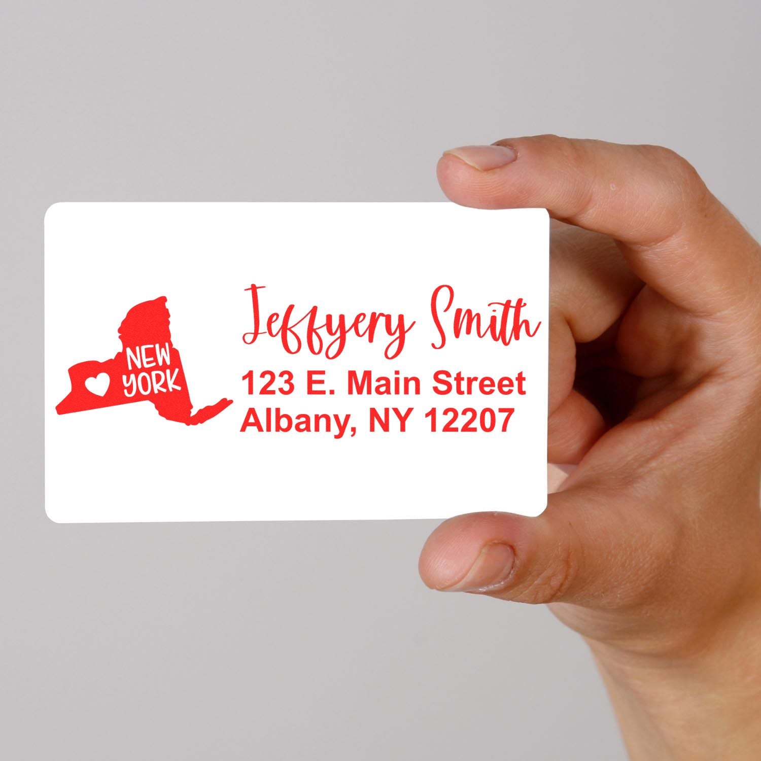 Hand holding a card with a red New York State Love Personalized Address Stamp design, featuring a heart and the text Jeffery Smith, 123 E. Main Street, Albany, NY 12207 in red.