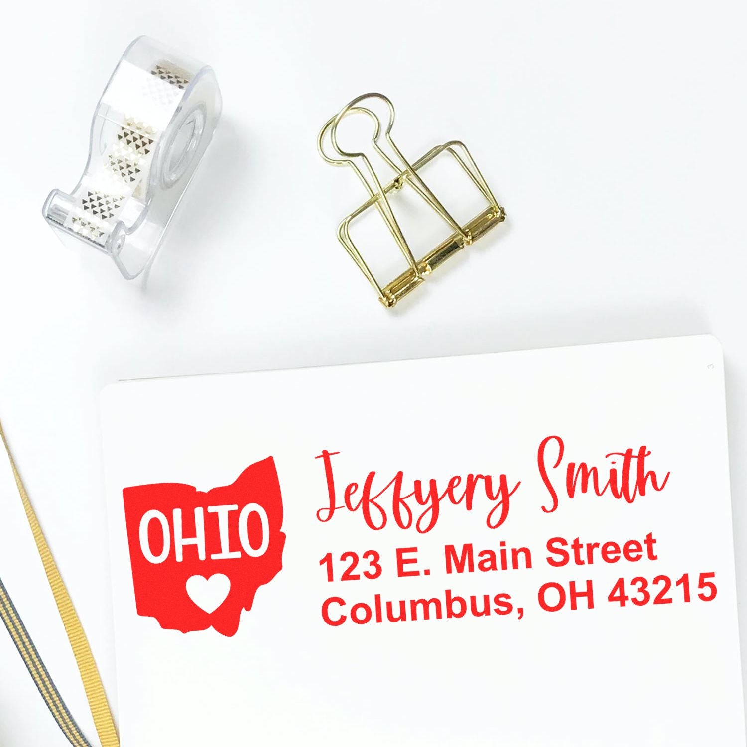 Ohio State Love Personalized Address Stamp on white paper with red text, featuring an Ohio map design. Nearby are a gold paper clip, tape dispenser, and ribbon.