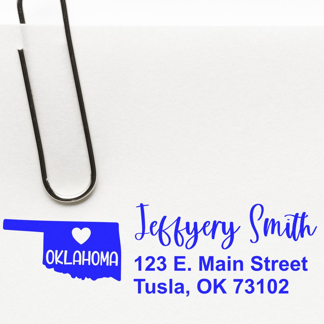 Slim Oklahoma Custom Address Stamp for Envelopes displayed on white paper with a paperclip. Blue ink shows a heart over Oklahoma, personalized with name and address.