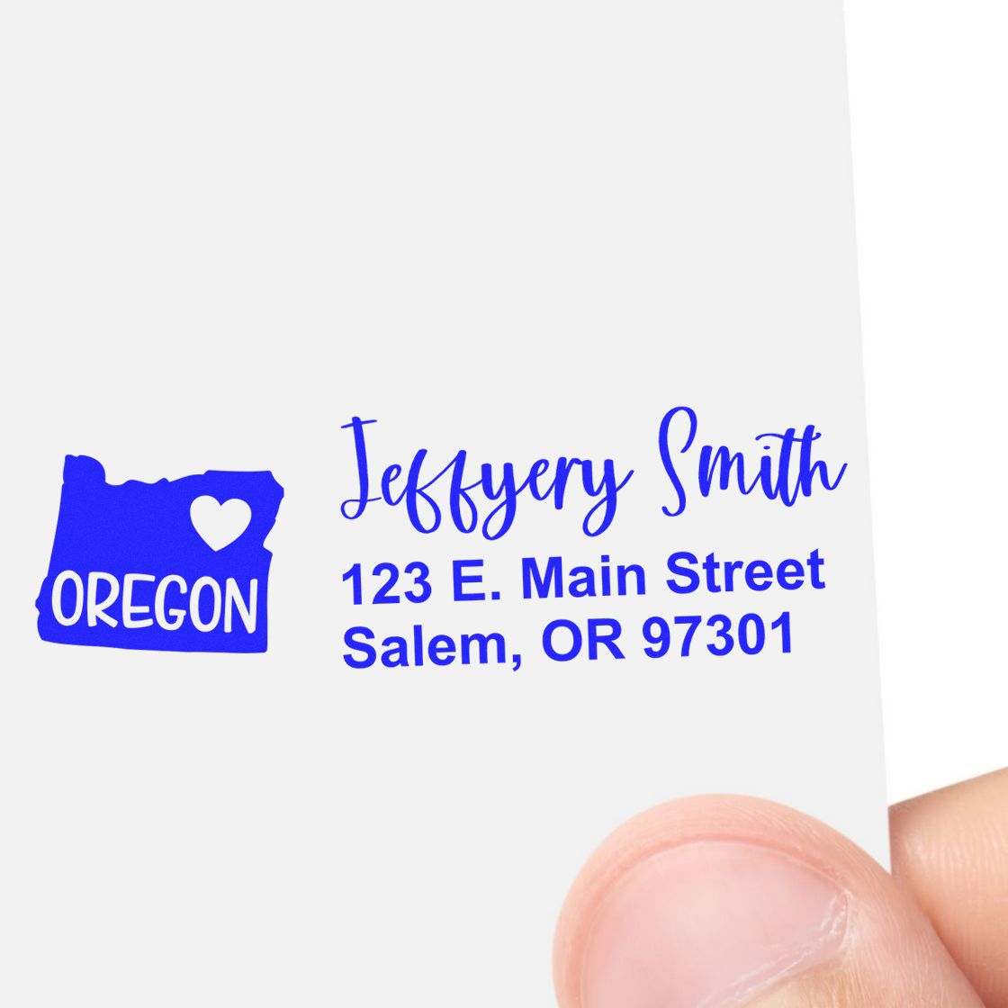 Close-up of a hand holding a card with the Oregon State Love Personalized Address Stamp, featuring a blue Oregon map with a heart and the address: Jeffery Smith, 123 E. Main Street, Salem, OR 97301.