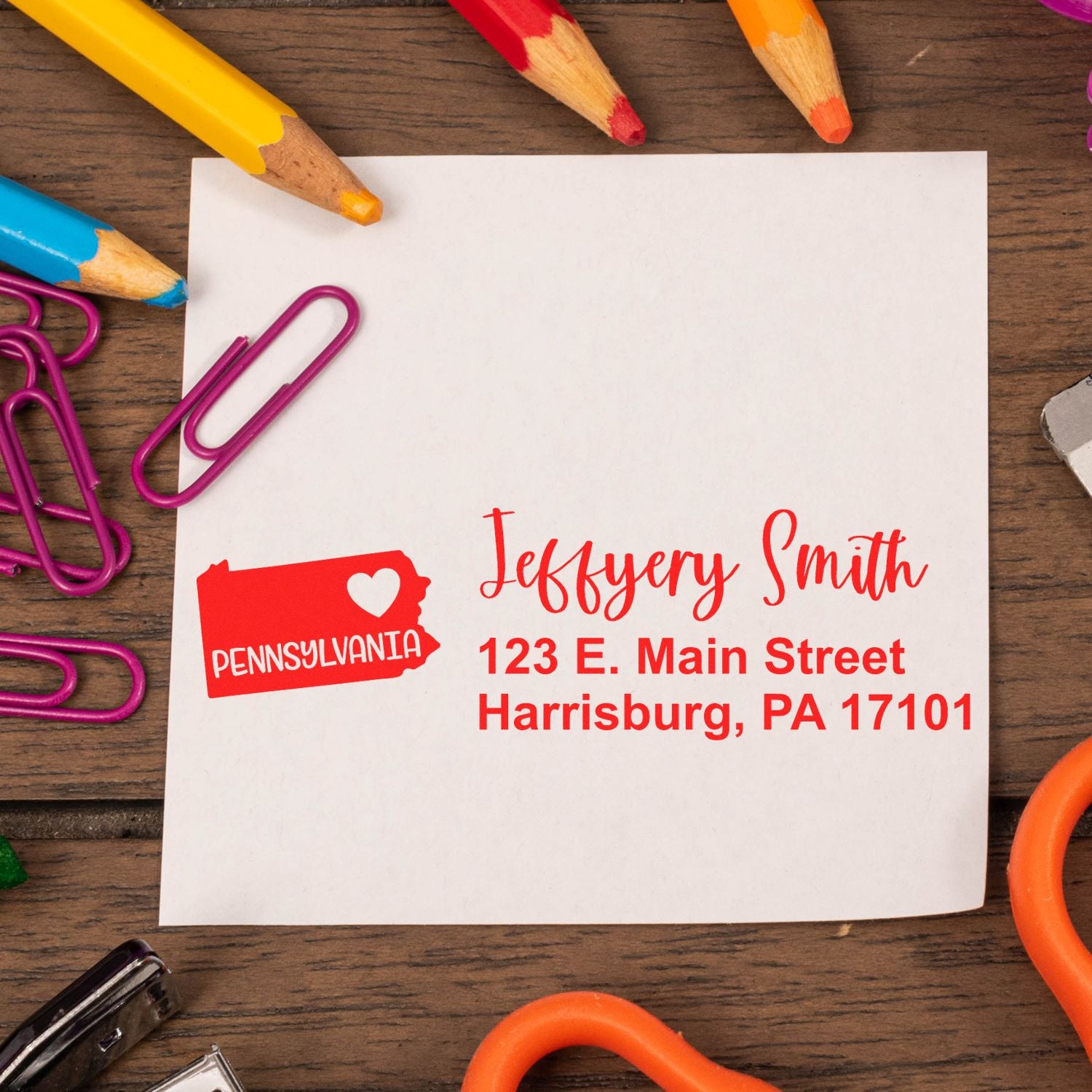 A personalized note with the Pennsylvania State Love Personalized Address Stamp, featuring a red state outline and heart, surrounded by colorful pencils and paperclips on a wooden desk.