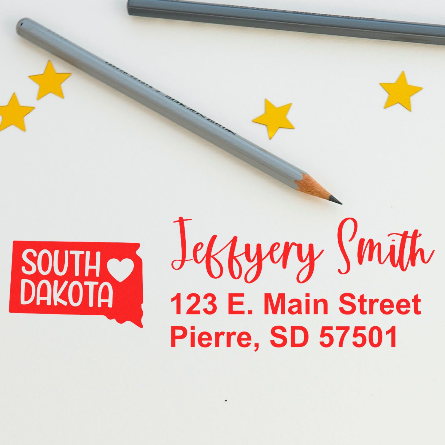 Image of the Slim South Dakota Custom Address Stamp for Envelopes, featuring a stamped address in red ink with a South Dakota outline, next to pencils and star stickers on a white surface.