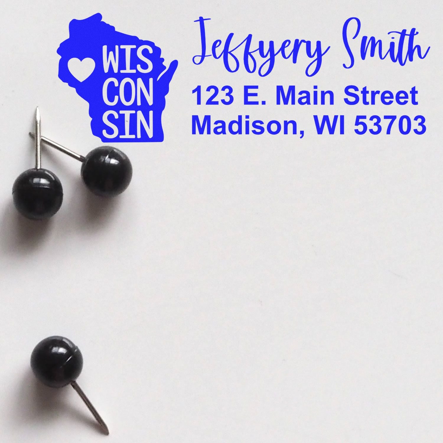 Slim Wisconsin Custom Address Stamp for Envelopes displayed with blue text and a Wisconsin state outline, featuring a sample address. Black push pins are placed nearby on a white background.