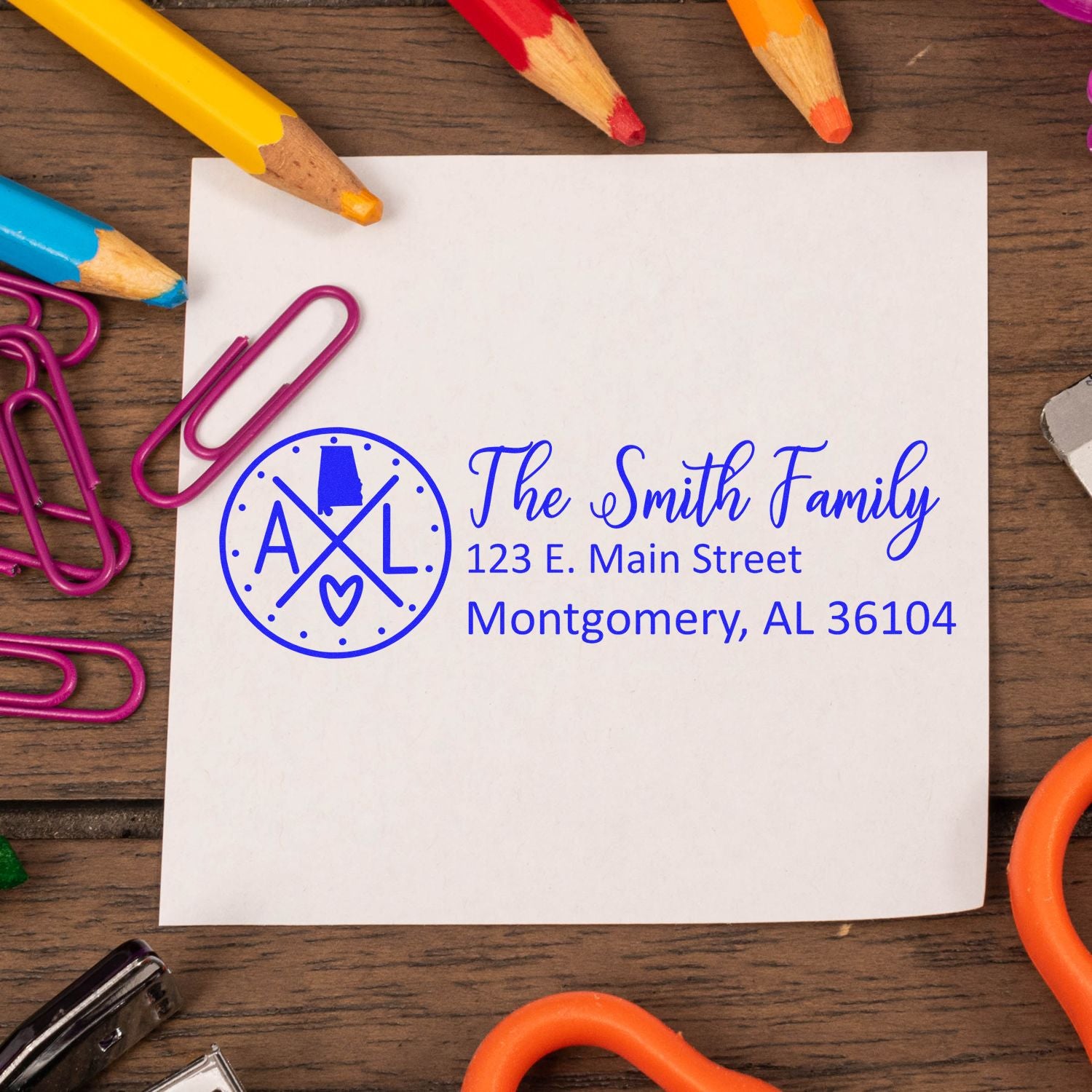 PSI Pre-Inked Alabama State Pride Customized Address Stamp on a white paper with colorful pencils and paperclips on a wooden surface.