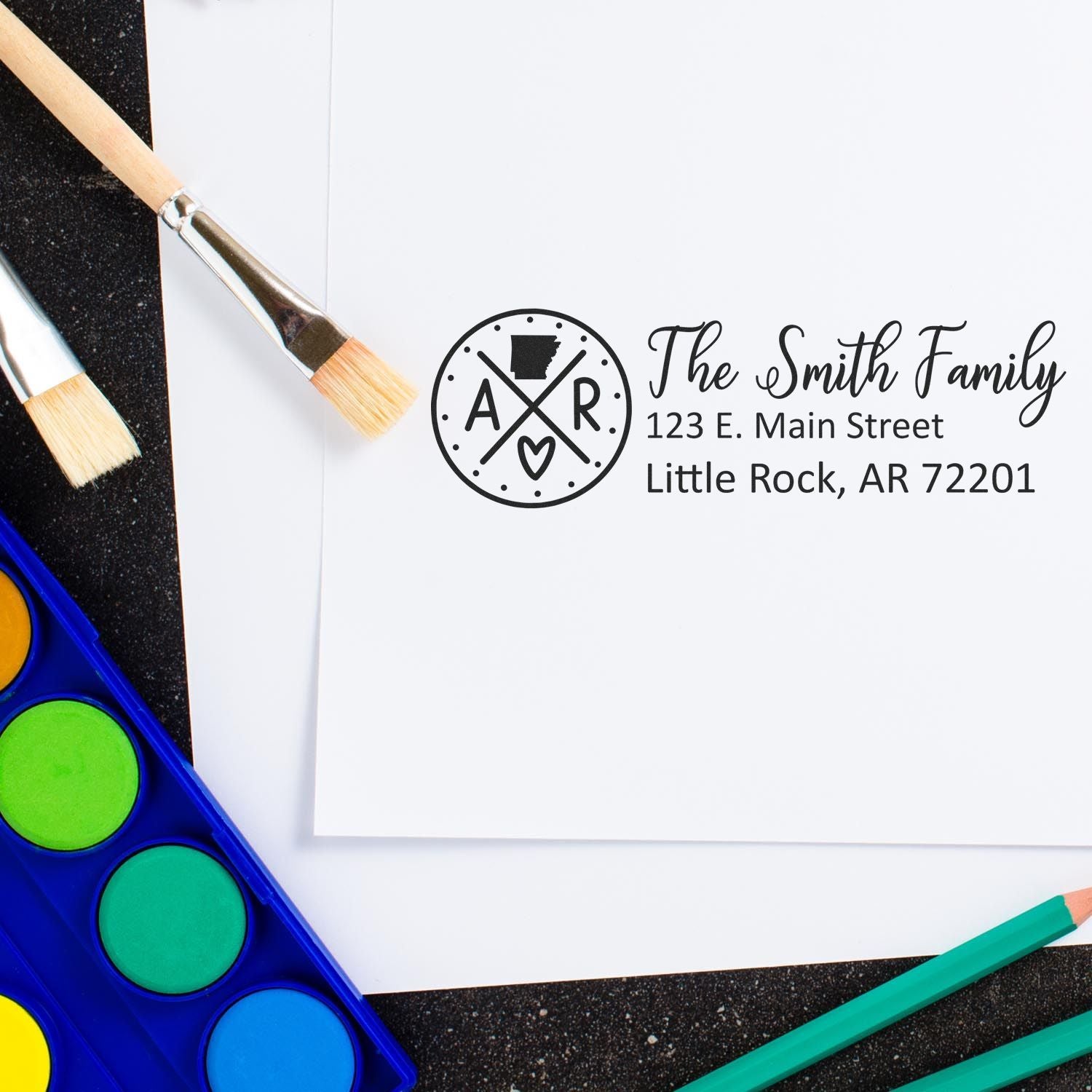 Self-Inking State Love of Arkansas Custom Address Stamp on white paper with paintbrushes and watercolor set nearby, featuring The Smith Family and an Arkansas design.