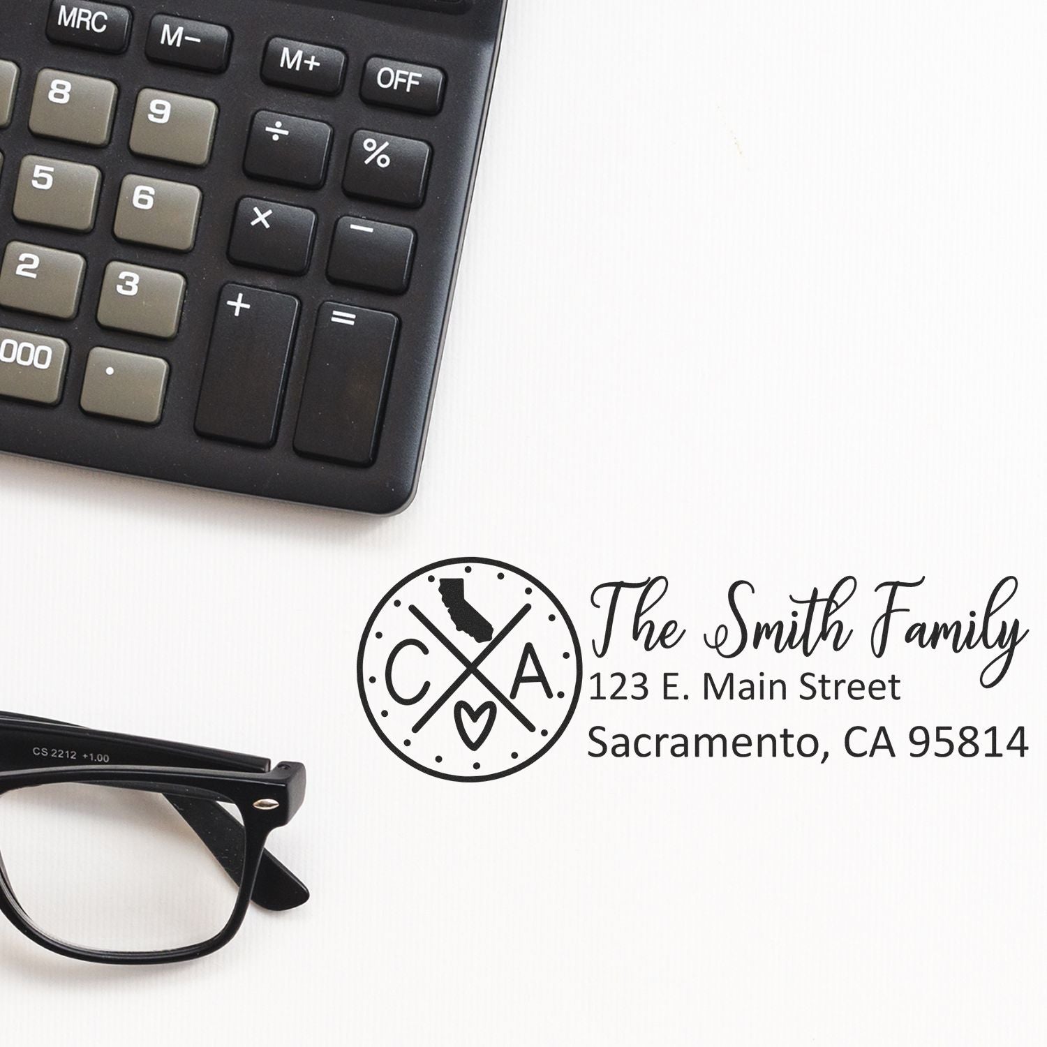 A Slim California Customized Pre-Inked Address Stamp on paper next to a calculator and glasses, displaying The Smith Family, 123 E. Main Street, Sacramento, CA 95814 in elegant script.