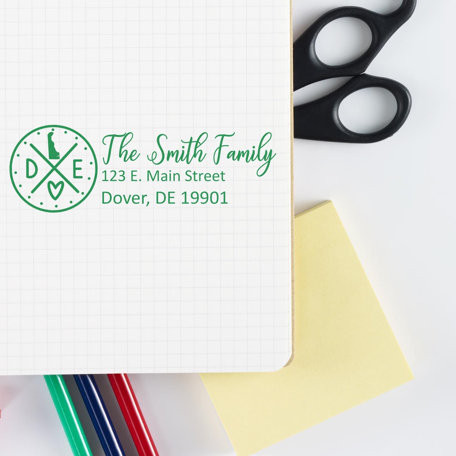 Slim Delaware Customized Pre-Inked Address Stamp displayed on a notebook with green ink, featuring a personalized address. Scissors, sticky notes, and colored pens are nearby on a white surface.