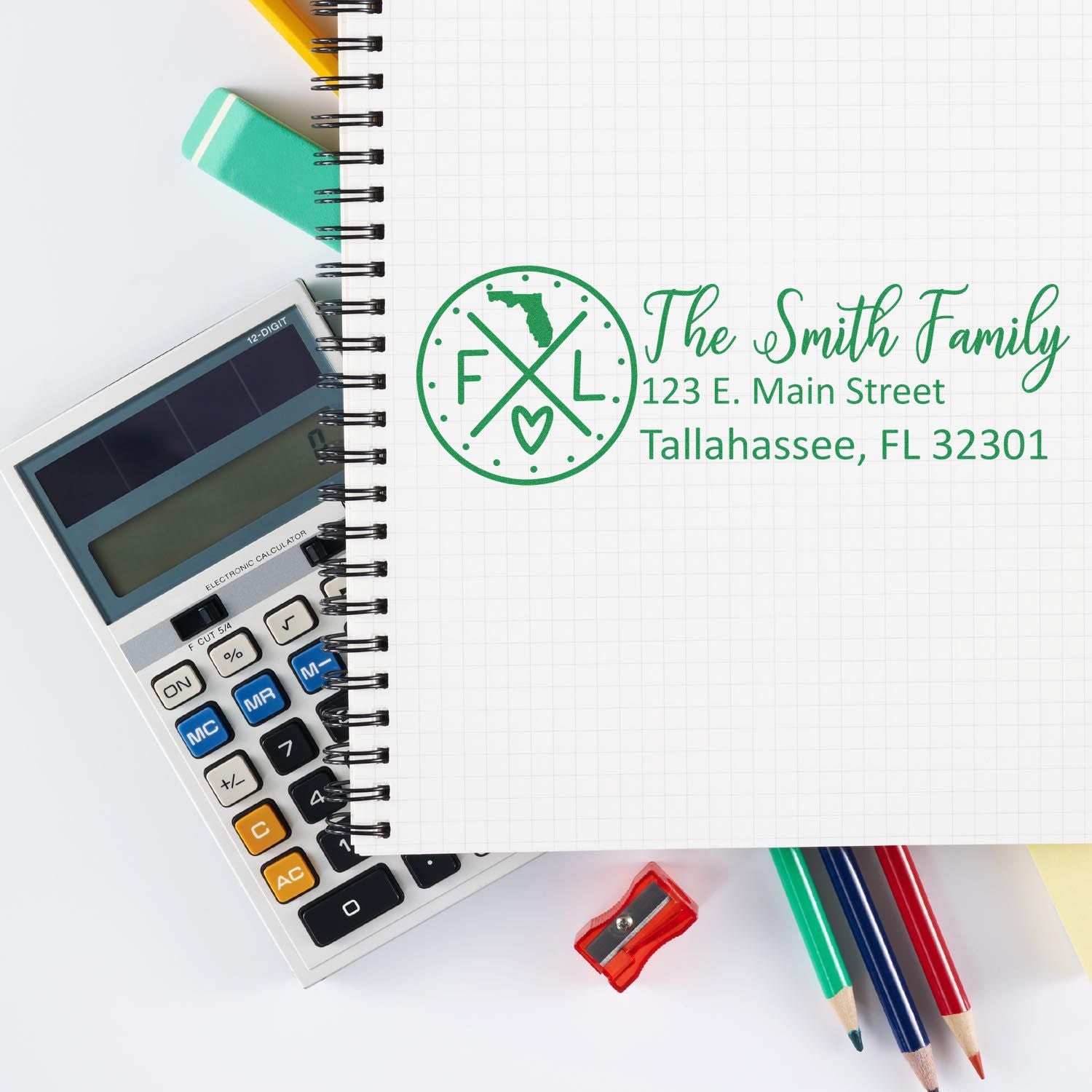 Slim Florida Customized Pre-Inked Address Stamp on a notebook page with a calculator and colorful pens, displaying The Smith Family, 123 E. Main Street, Tallahassee, FL 32301 in green ink.