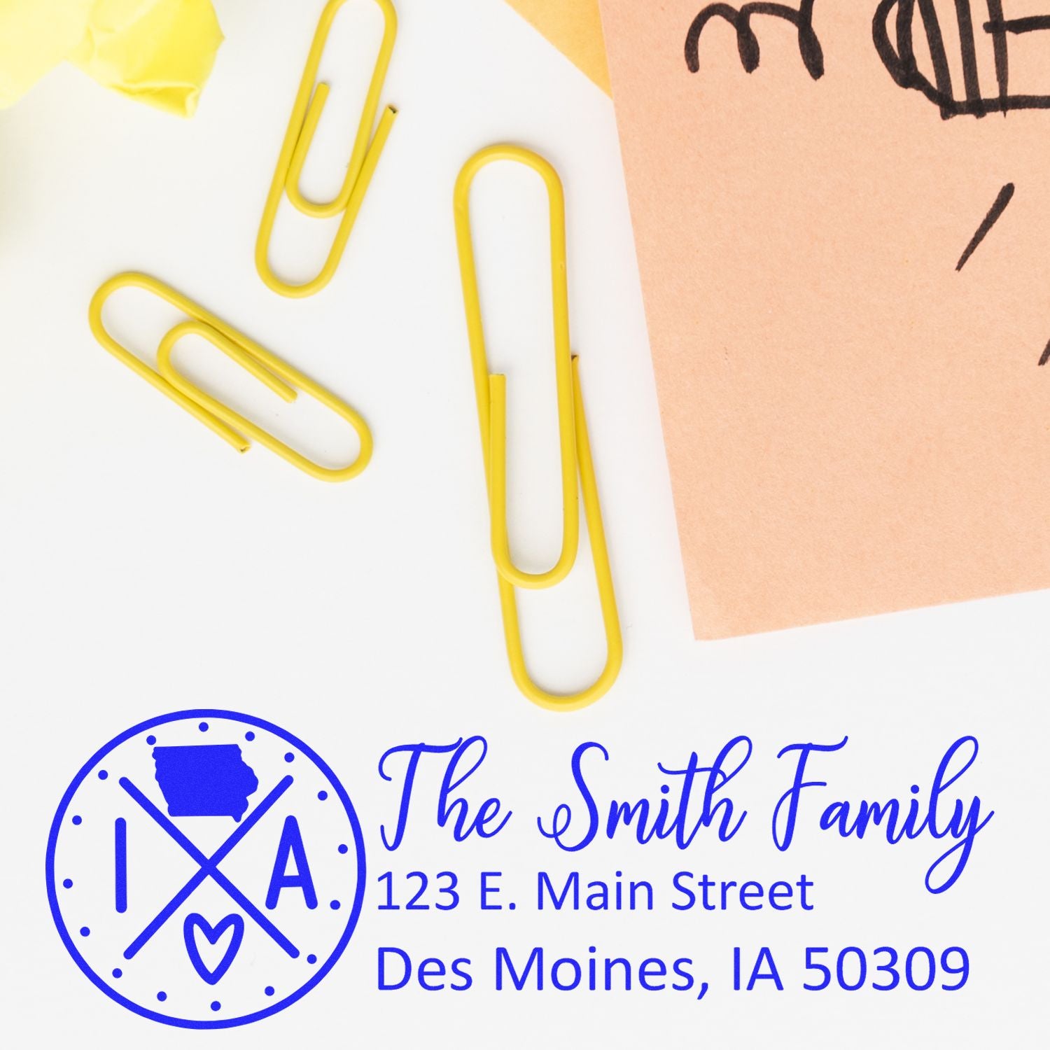 Yellow paperclips and a pink paper on a desk with the Self-Inking State Love of Iowa Custom Address Stamp imprint showing The Smith Family, 123 E. Main Street, Des Moines, IA 50309 in blue.