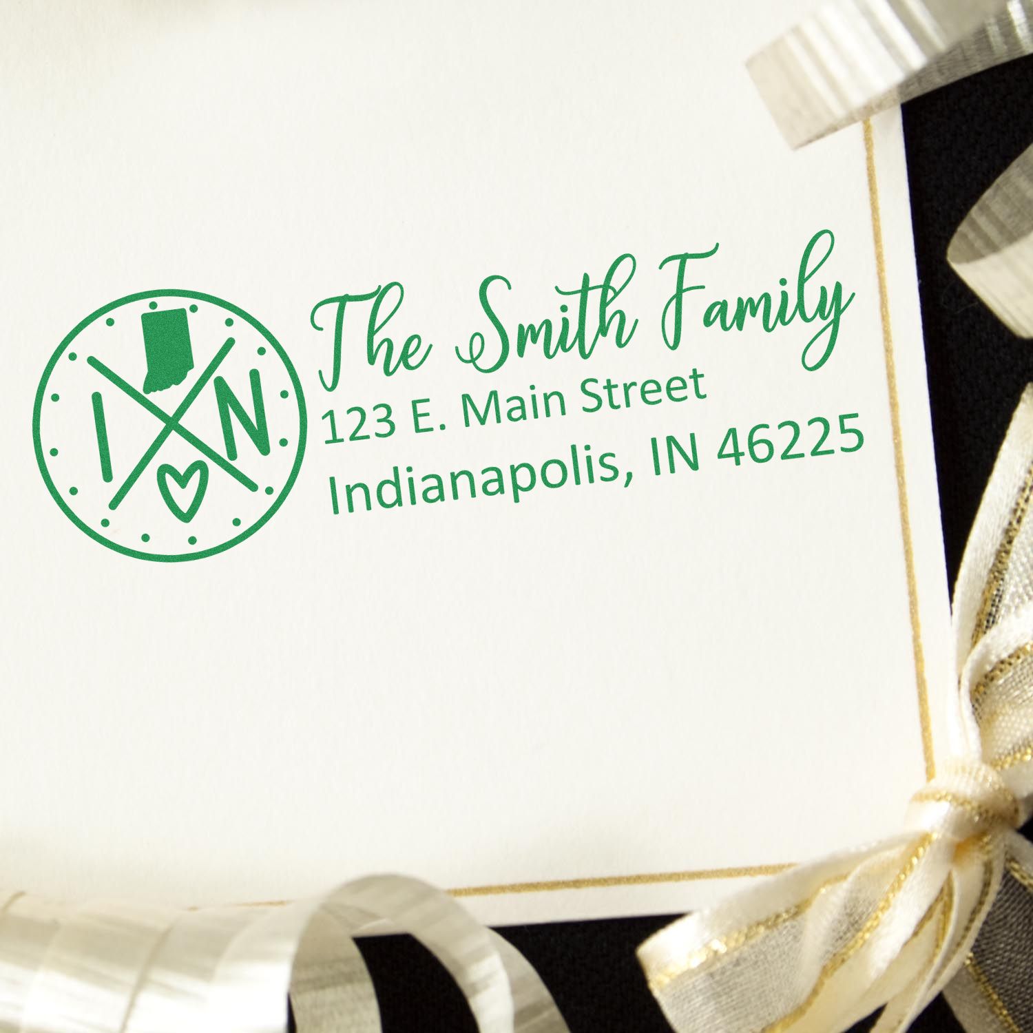 Slim Indiana Customized Pre-Inked Address Stamp on an envelope with a green design, featuring a family name and address. The envelope is adorned with a gold and black ribbon.