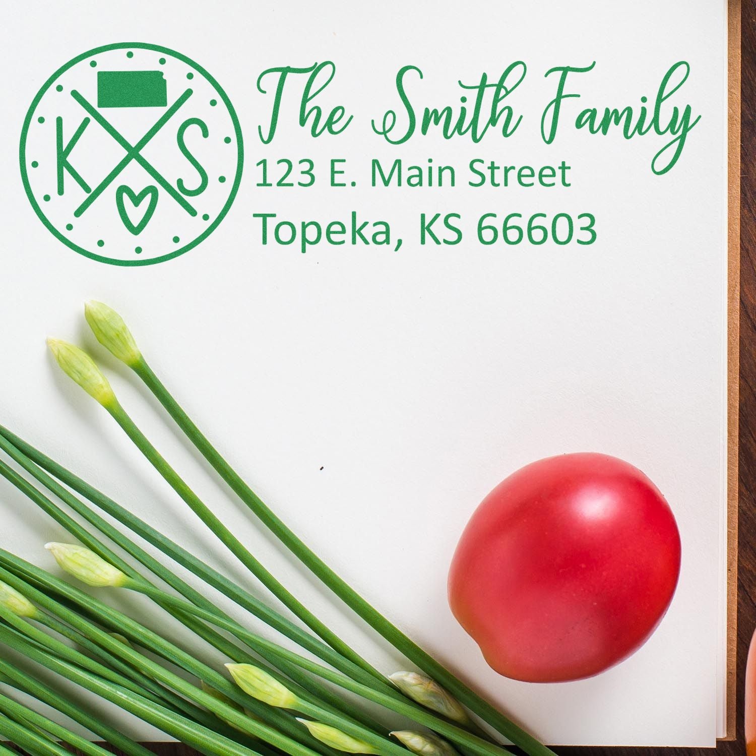 A PSI Pre-Inked Kansas State Pride Customized Address Stamp on paper with The Smith Family address in green ink, next to green onions and a red tomato on a wooden surface.