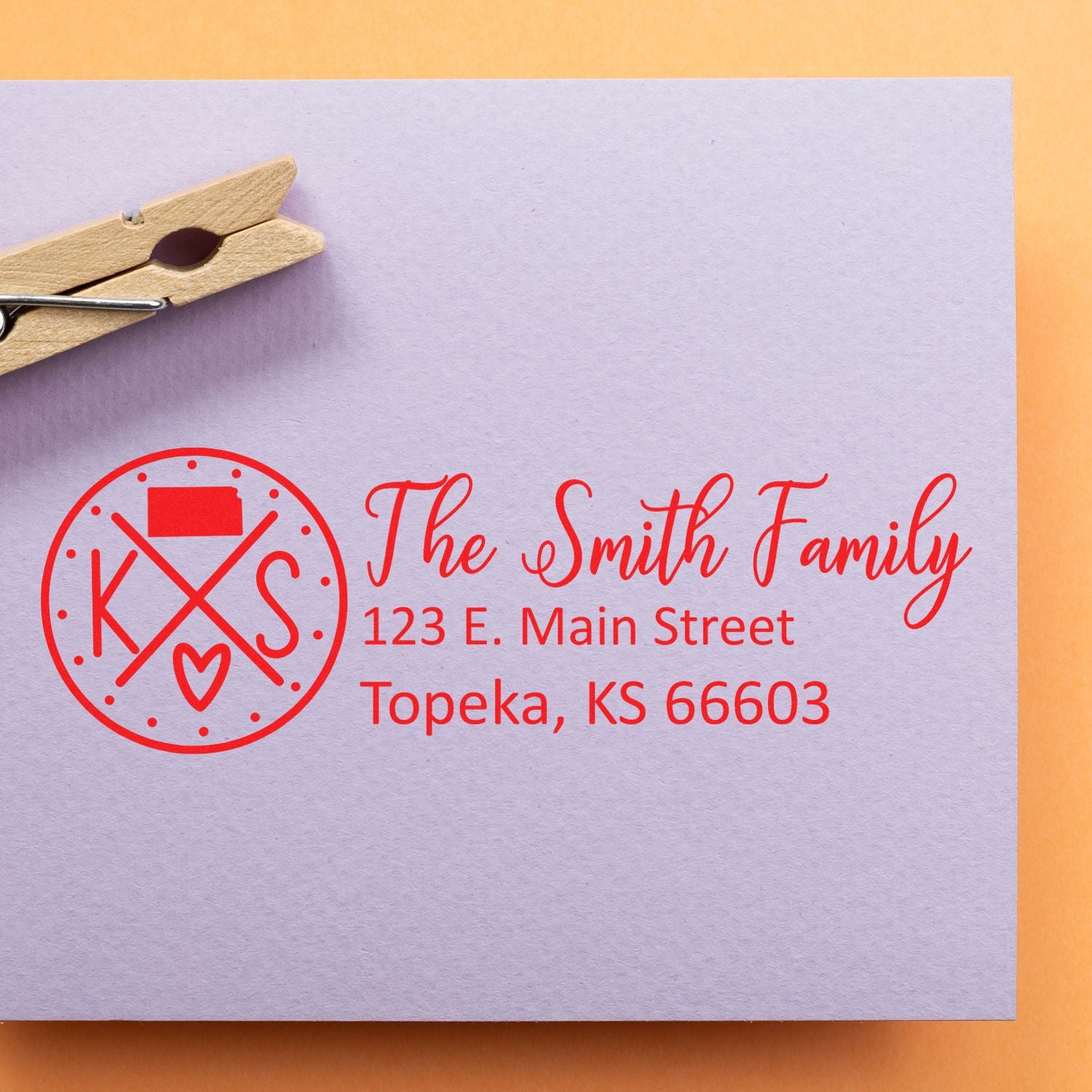 Slim Kansas Customized Pre-Inked Address Stamp on a lavender envelope with red ink, displaying The Smith Family, 123 E. Main Street, Topeka, KS 66603, next to a wooden clothespin.