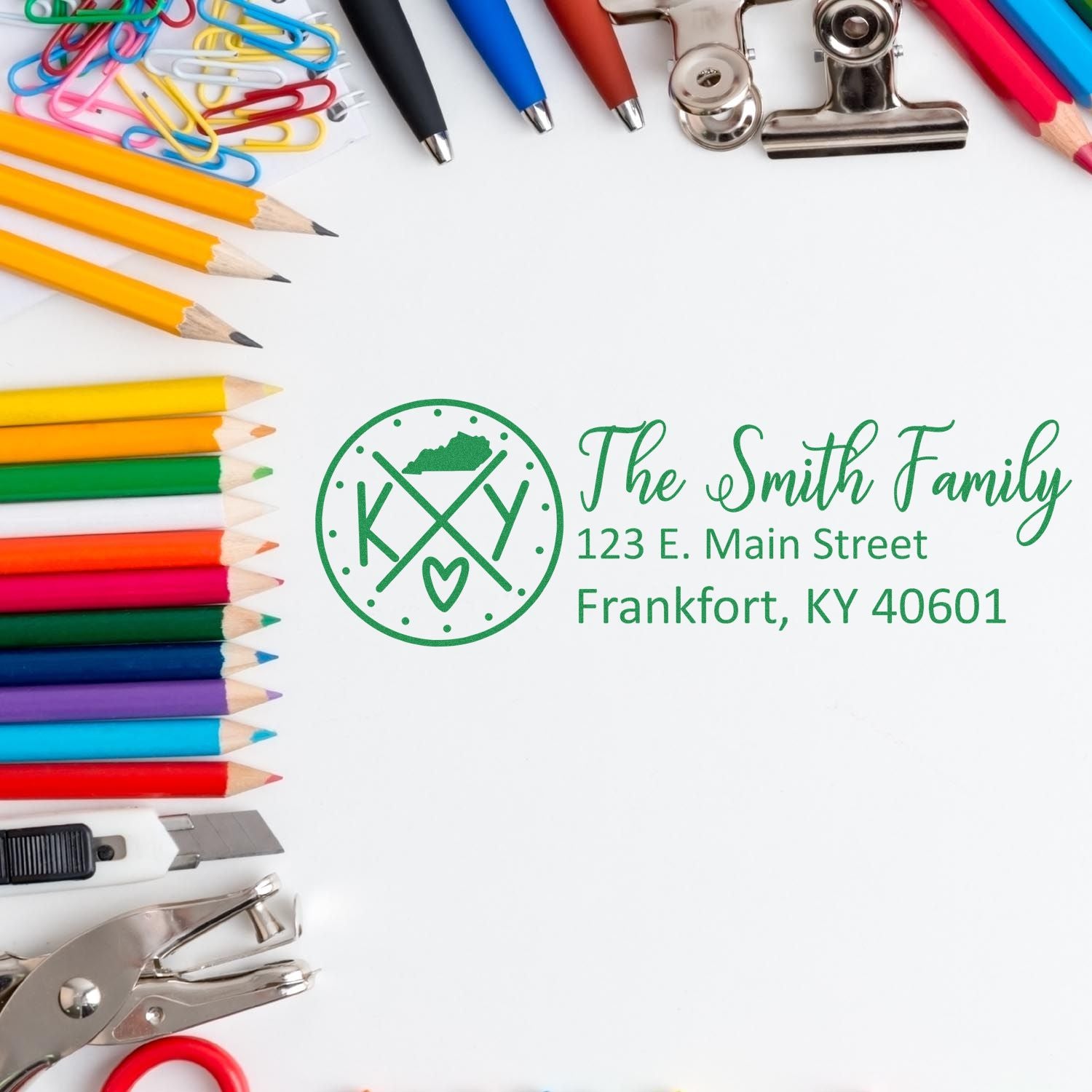 PSI Pre-Inked Kentucky State Pride Customized Address Stamp displayed on paper, surrounded by colorful pencils, paperclips, and office supplies. Stamp shows The Smith Family with a Kentucky state design.