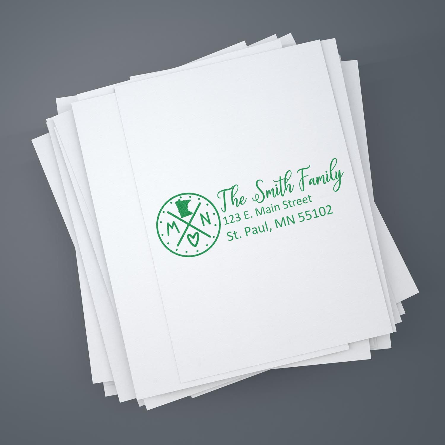 Stack of white papers stamped with green ink using the Slim Minnesota Customized Pre-Inked Address Stamp, featuring a circular MN logo and address for The Smith Family in St. Paul, MN.