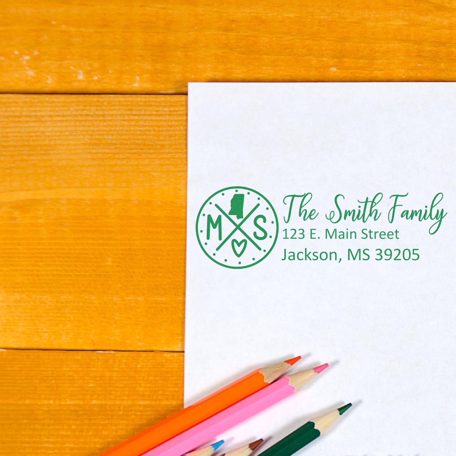 Mississippi State Pride Customized Address Stamp on white paper with The Smith Family and address in green ink, surrounded by colorful pencils on a wooden surface.