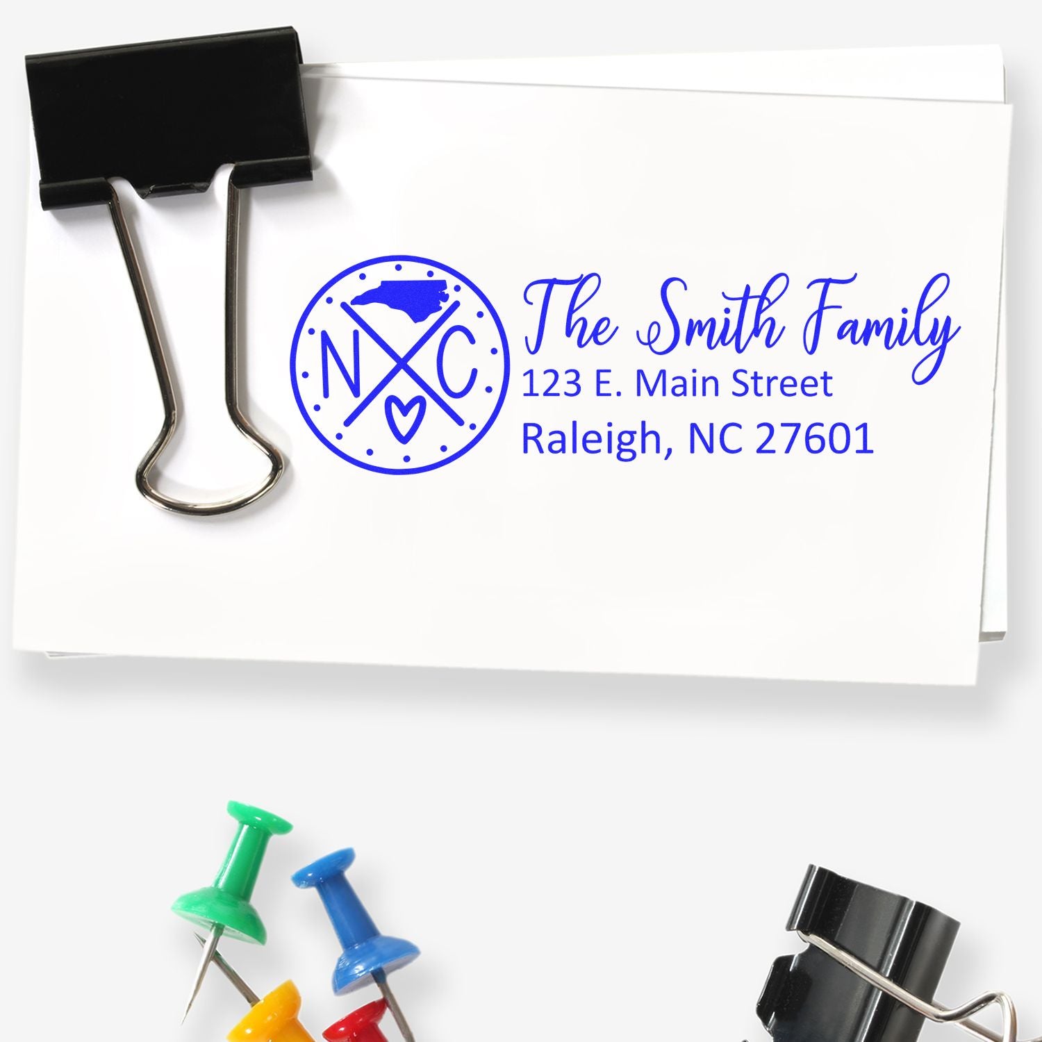 Slim North Carolina Customized Pre-Inked Address Stamp on envelope with paper clips and push pins. Features 'The Smith Family' and Raleigh address in blue ink.