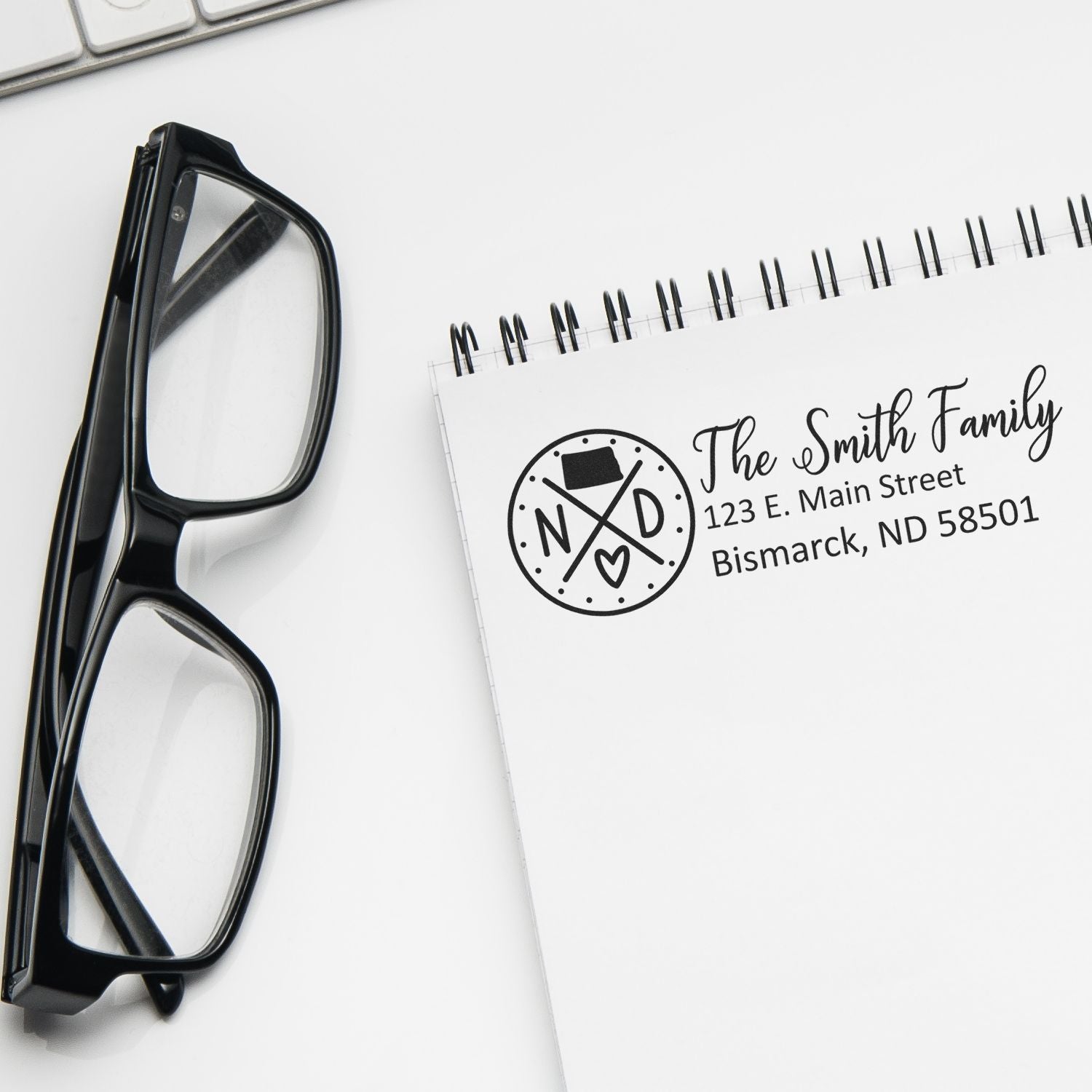 A notebook with a stamped address reading The Smith Family, 123 E. Main Street, Bismarck, ND 58501 using the Self-Inking State Love of North Dakota Custom Address Stamp, next to a pair of glasses.