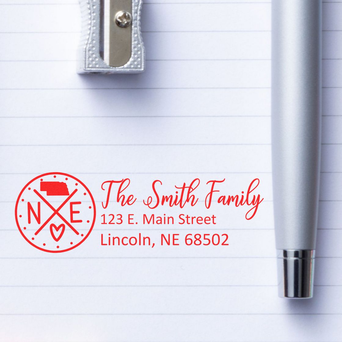 Nebraska State Pride Customized Address Stamp featuring a red design with The Smith Family and an address. Displayed on lined paper with a silver pen and pencil sharpener nearby.