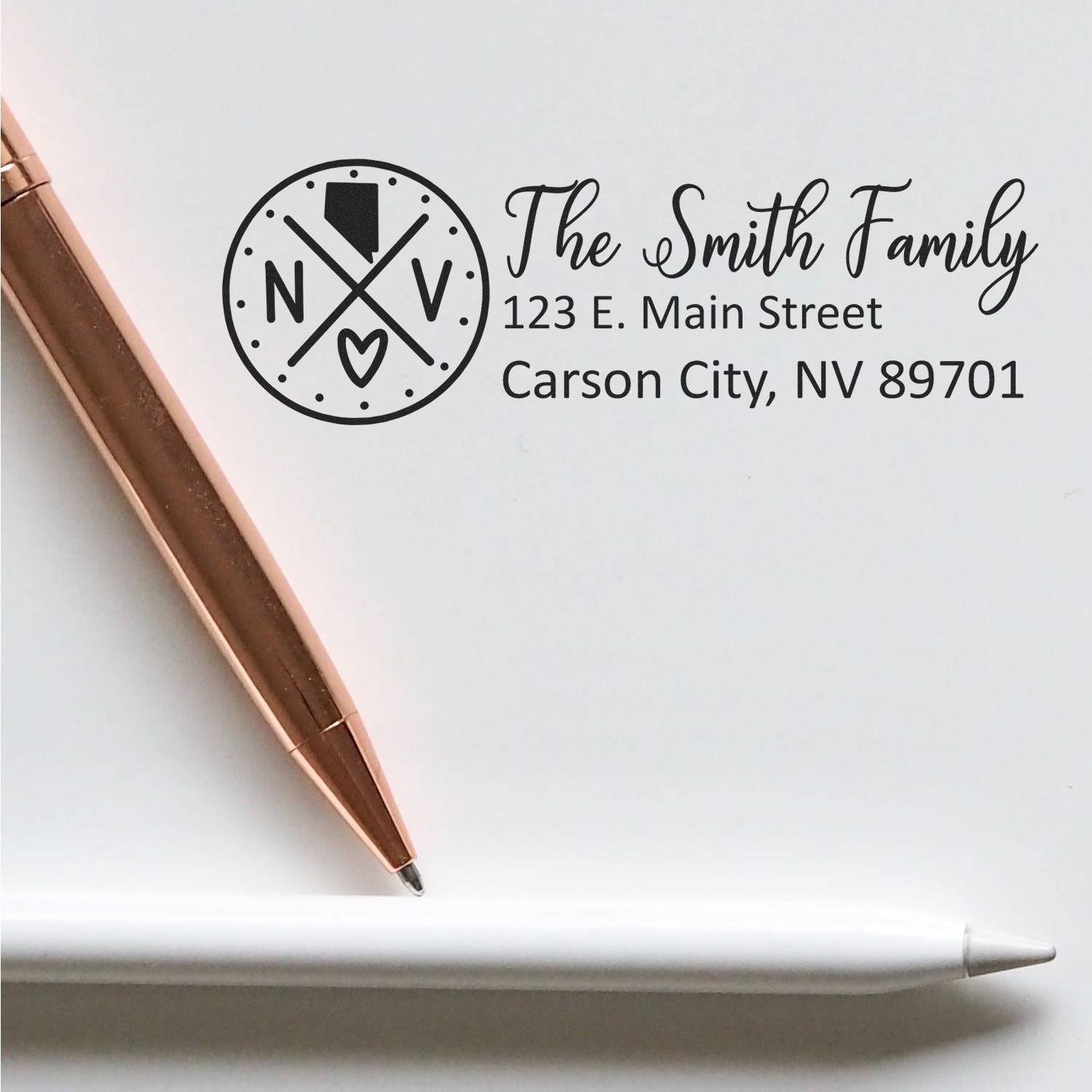 A Slim Nevada Customized Pre-Inked Address Stamp is shown on paper with a rose gold pen and white stylus, featuring a personalized address for The Smith Family in Carson City, NV.