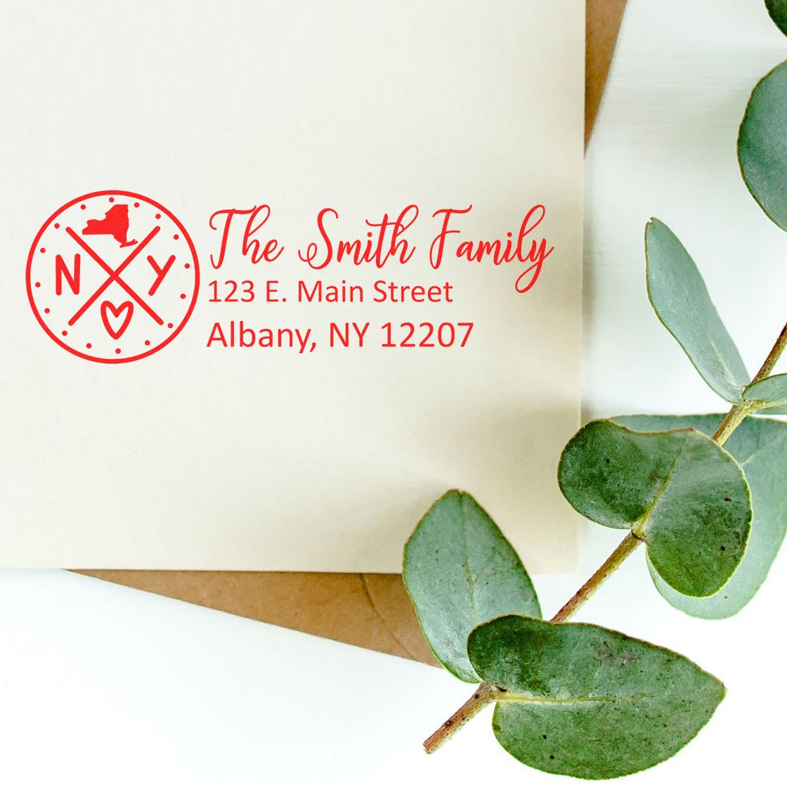 A Slim New York Customized Pre-Inked Address Stamp marks an envelope with a red design featuring The Smith Family and an address, next to green eucalyptus leaves on a white background.