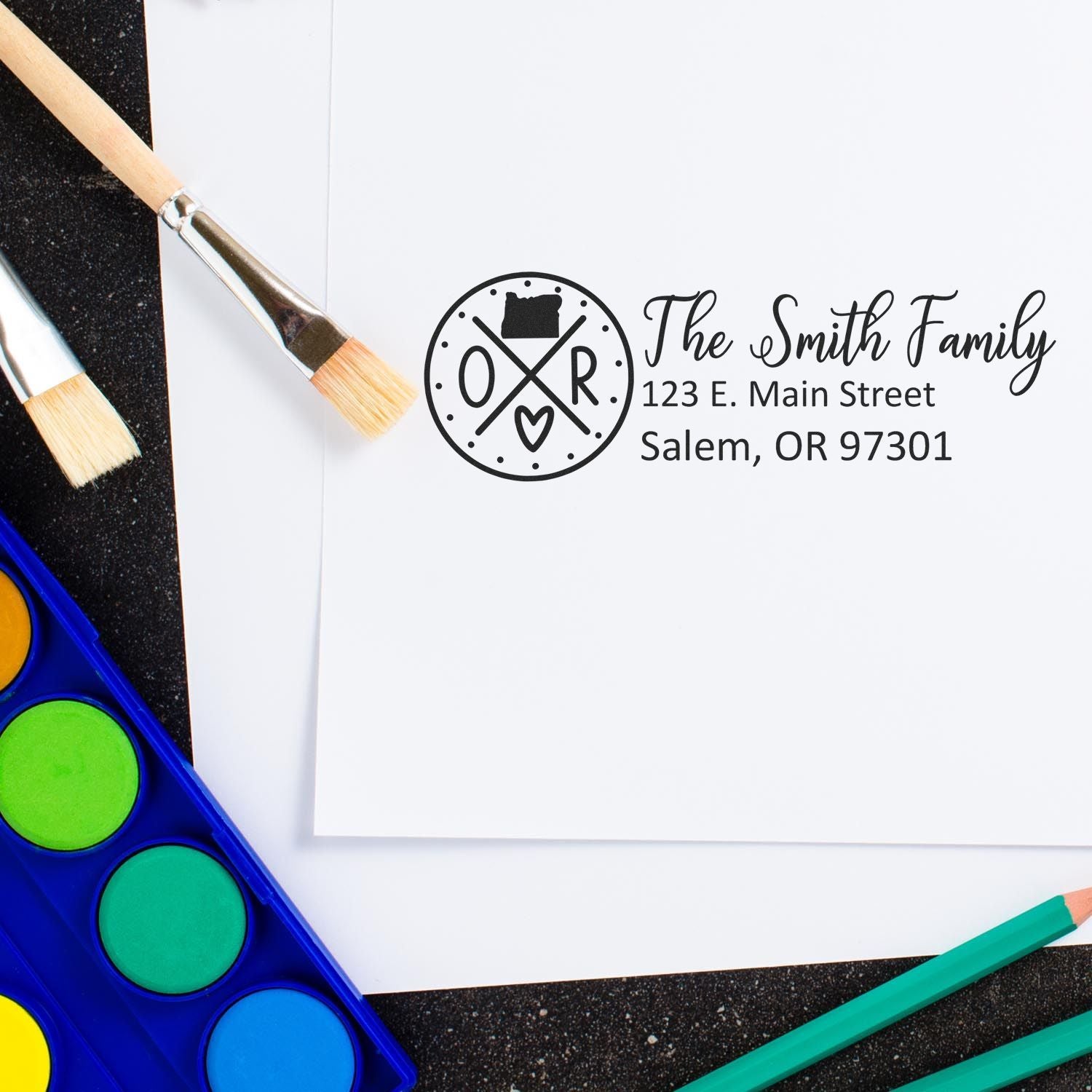 Slim Oregon Customized Pre-Inked Address Stamp on white paper with The Smith Family address. Surrounded by paintbrushes and watercolor paints on a black surface.