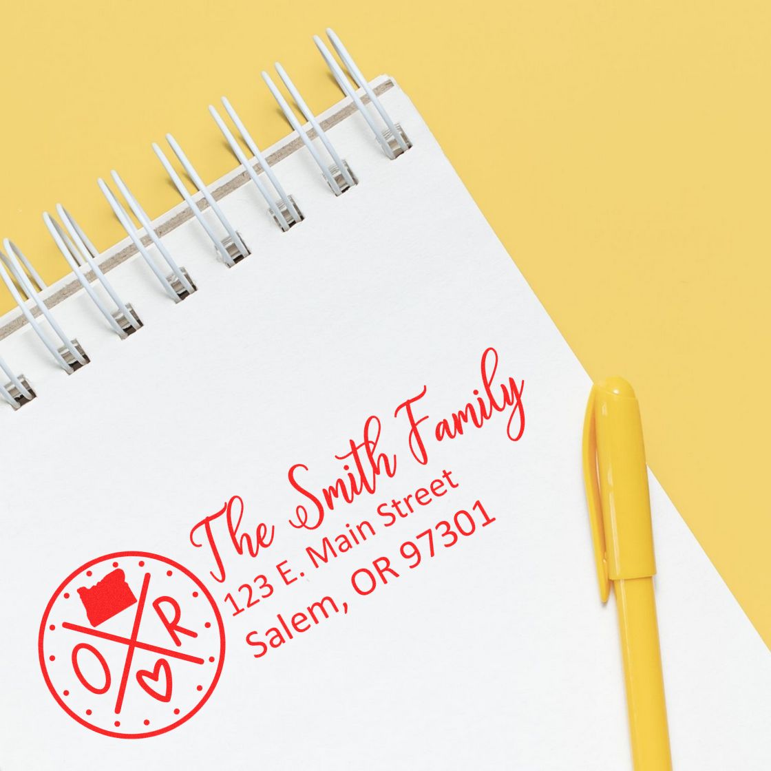 Self-Inking State Love of Oregon Custom Address Stamp on a notepad with red ink, displaying The Smith Family, 123 E. Main Street, Salem, OR 97301 beside a yellow pen on a yellow background.