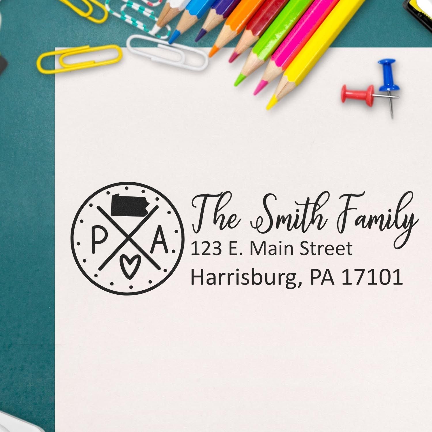 Slim Pennsylvania Customized Pre-Inked Address Stamp on white paper with colorful pencils, paperclips, and a pushpin nearby. The stamp displays a family name and address in a stylish design.