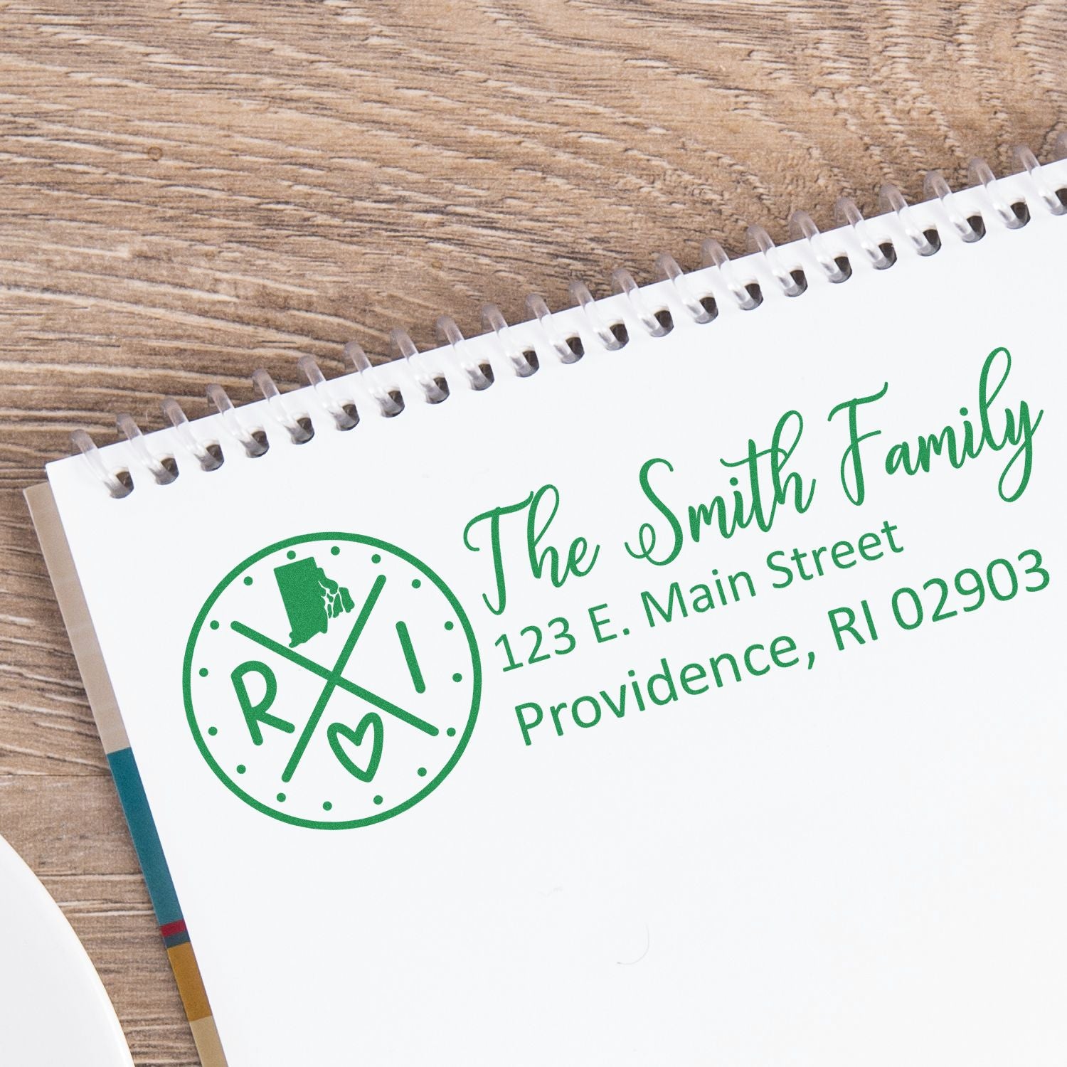 Slim Rhode Island Customized Pre-Inked Address Stamp on a notebook, displaying The Smith Family, 123 E. Main Street, Providence, RI 02903 in green ink with a decorative design.