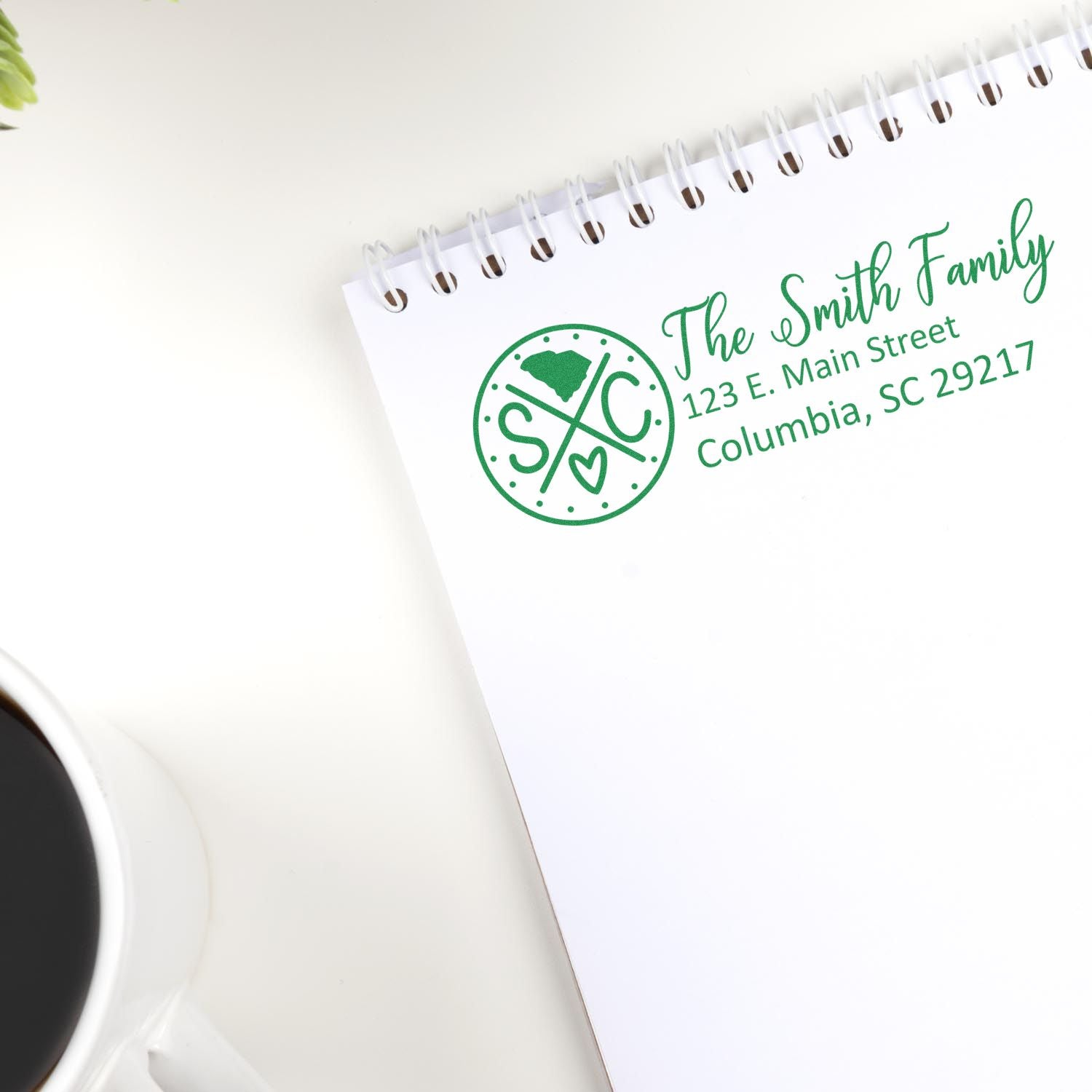 Slim South Carolina Customized Pre-Inked Address Stamp on a notepad with The Smith Family, 123 E. Main Street, Columbia, SC 29217 in green ink, next to a cup of coffee and a plant.