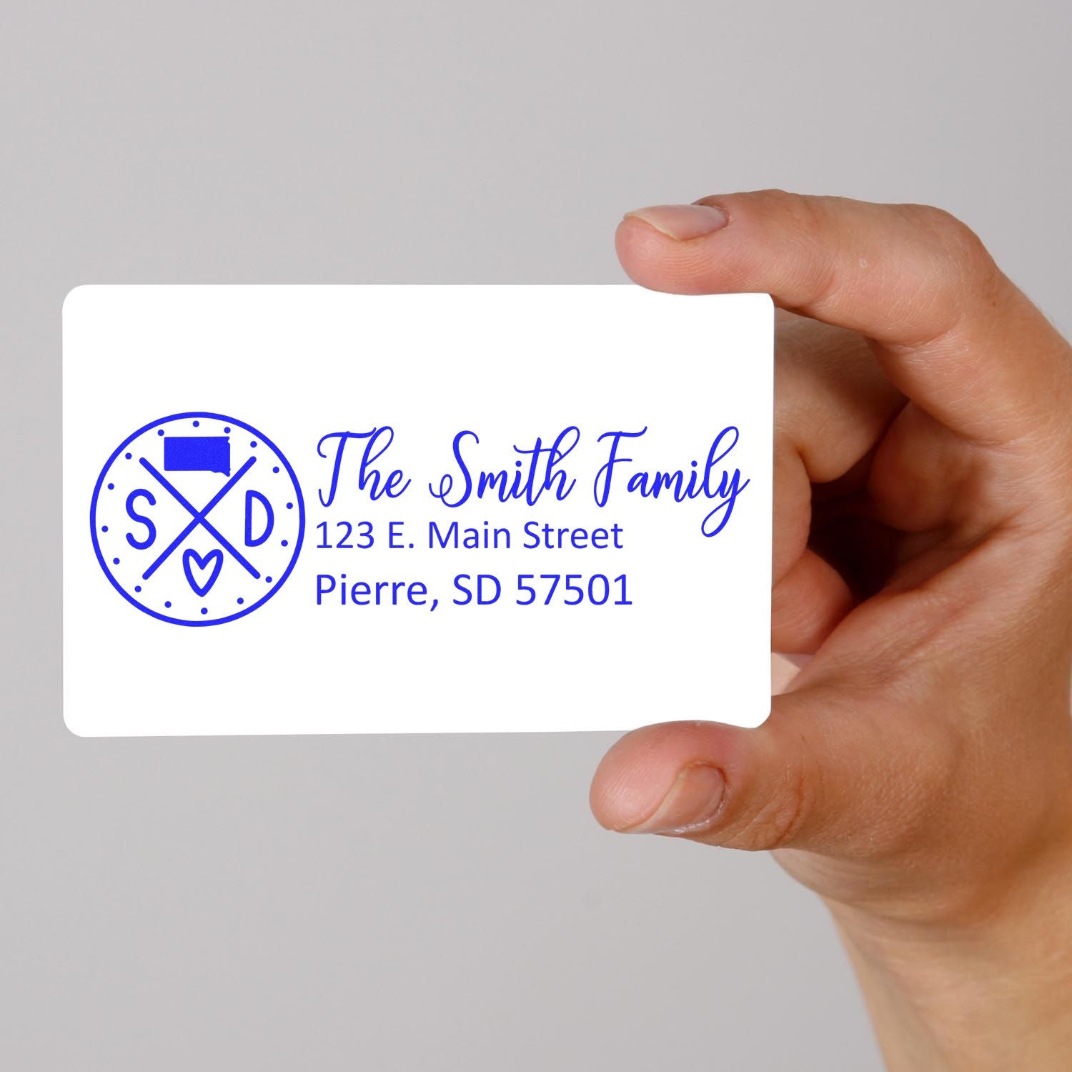 Hand holding a card with a PSI Pre-Inked South Dakota State Pride Customized Address Stamp design, featuring The Smith Family and an address in Pierre, SD, with a blue state-themed emblem.