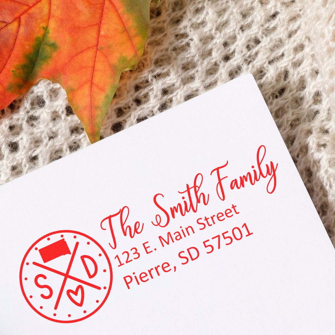 Self-Inking State Love of South Dakota Custom Address Stamp on a white envelope with red ink, featuring a state-themed design. Placed on a textured surface with an autumn leaf nearby.