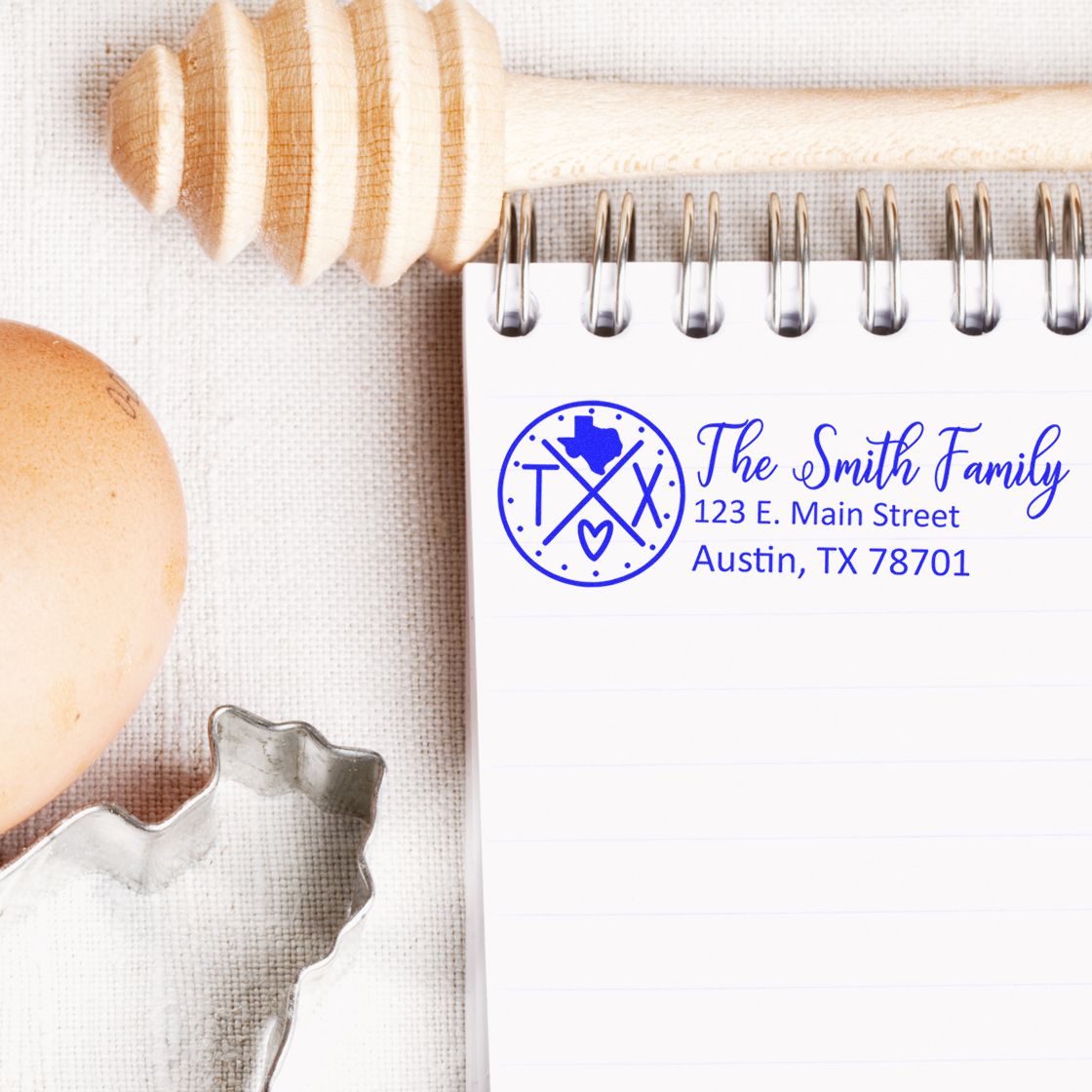 Slim Texas Customized Pre-Inked Address Stamp on a notepad with a honey dipper, egg, and cookie cutter nearby, displaying The Smith Family, 123 E. Main Street, Austin, TX 78701 in blue ink.