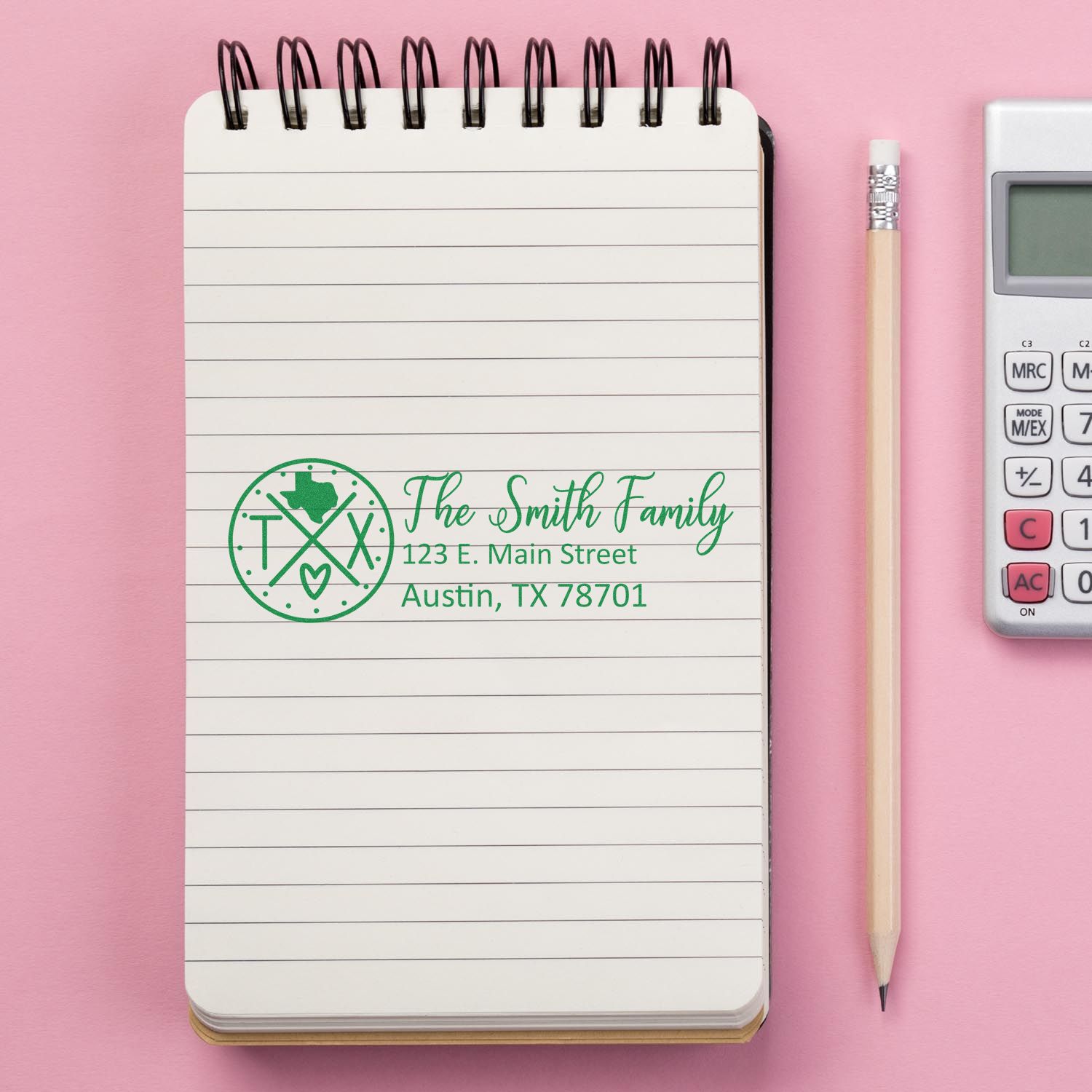 Slim Texas Customized Pre-Inked Address Stamp on a notepad with a pink background, next to a pencil and calculator. The stamp displays a family name and address in green ink.