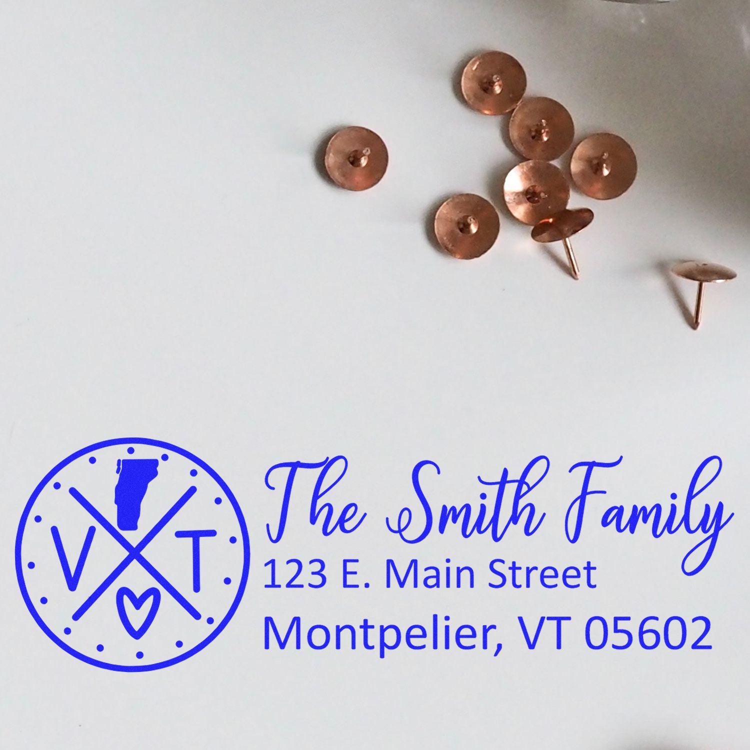 PSI Pre-Inked Vermont State Pride Customized Address Stamp with The Smith Family and Montpelier, VT address in blue ink, next to copper tacks on a white surface.