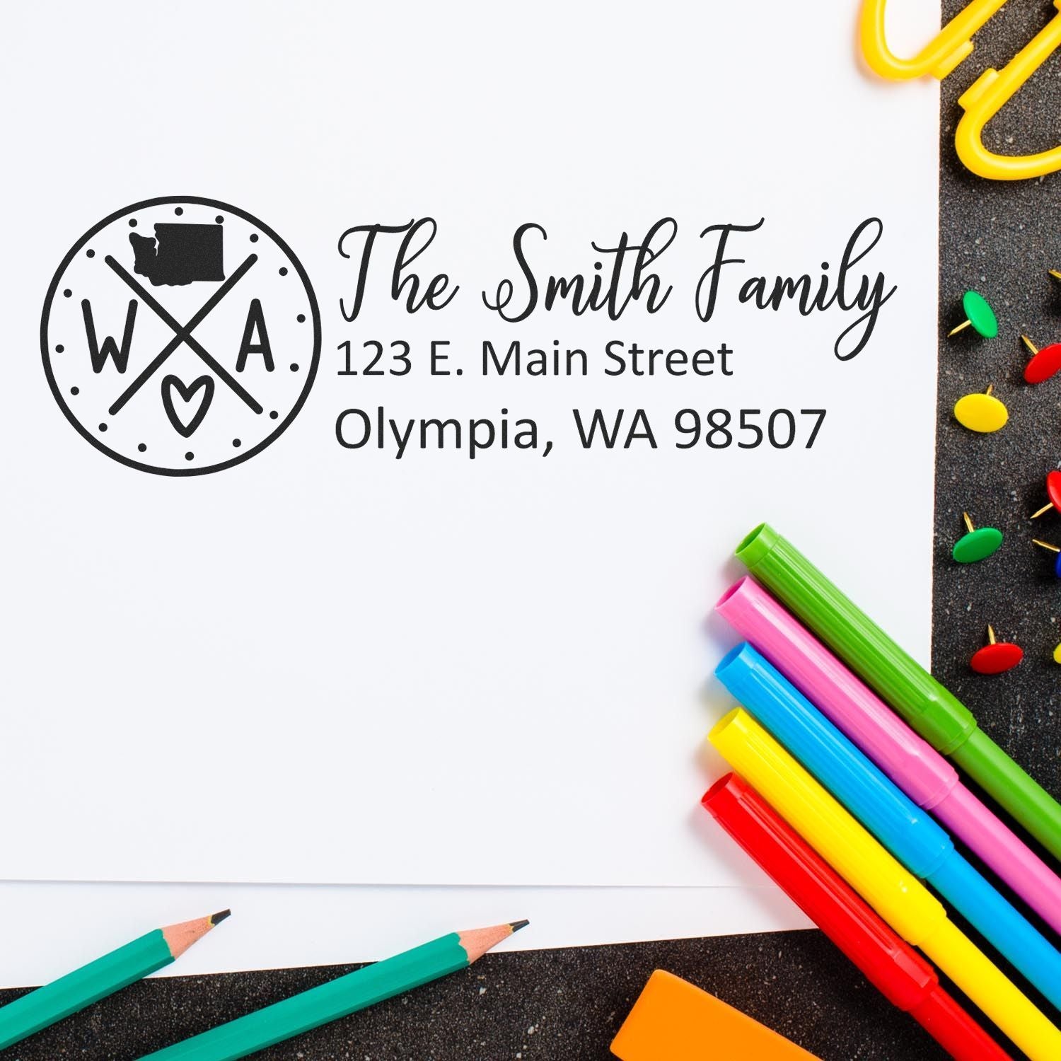 Slim Washington Customized Pre-Inked Address Stamp on white paper with colorful pens and push pins. The stamp displays The Smith Family, 123 E. Main Street, Olympia, WA 98507 with a decorative design.