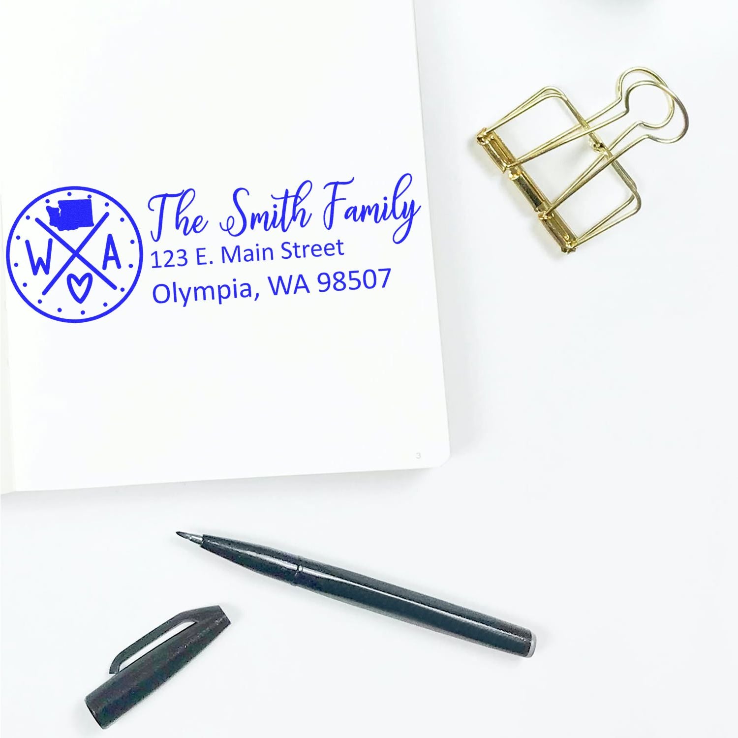 Slim Washington Customized Pre-Inked Address Stamp on white paper with The Smith Family address in blue ink, next to a black pen and gold binder clip on a white surface.