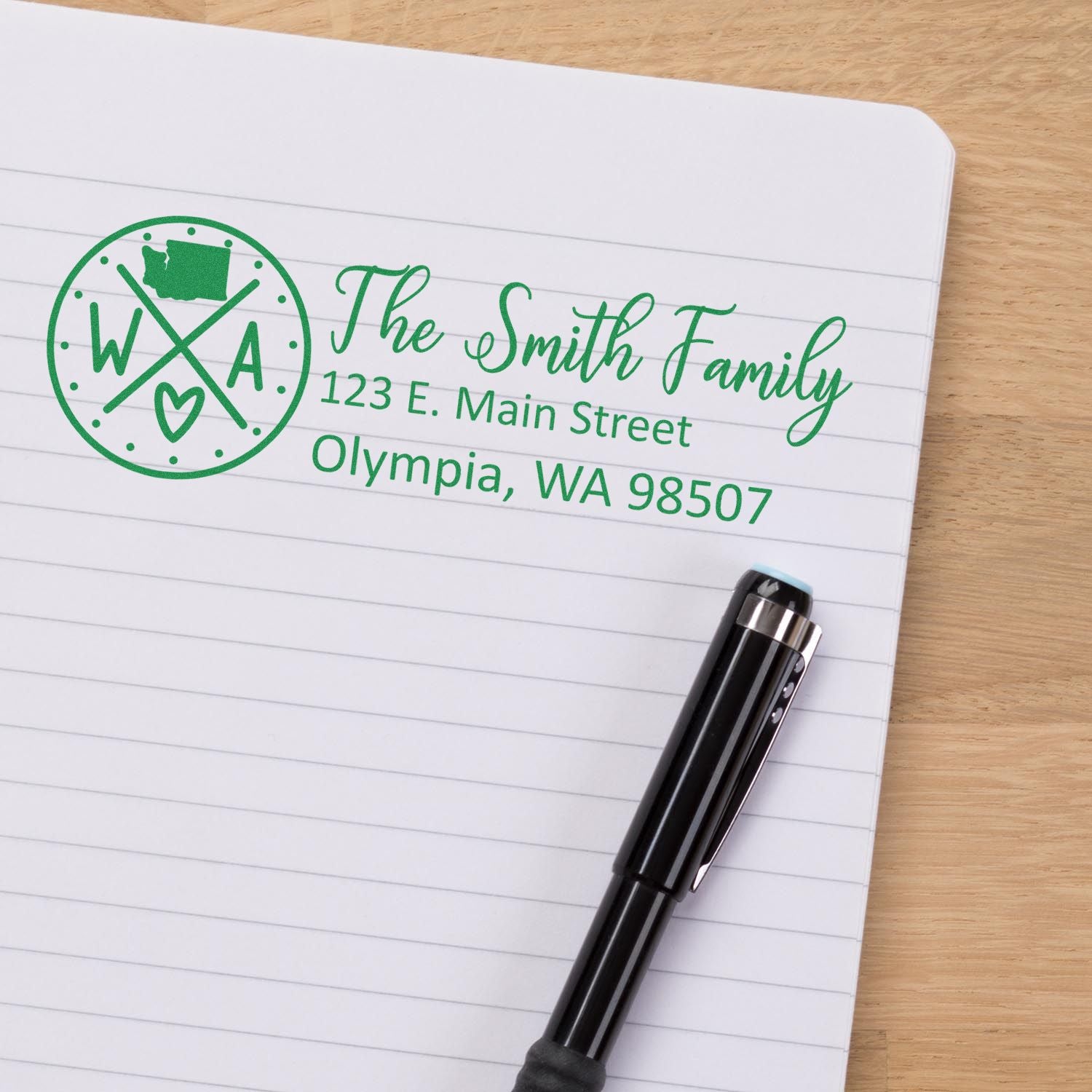 Slim Washington Customized Pre-Inked Address Stamp on paper, displaying The Smith Family, 123 E. Main Street, Olympia, WA 98507 in green ink, with a pen nearby on a wooden surface.