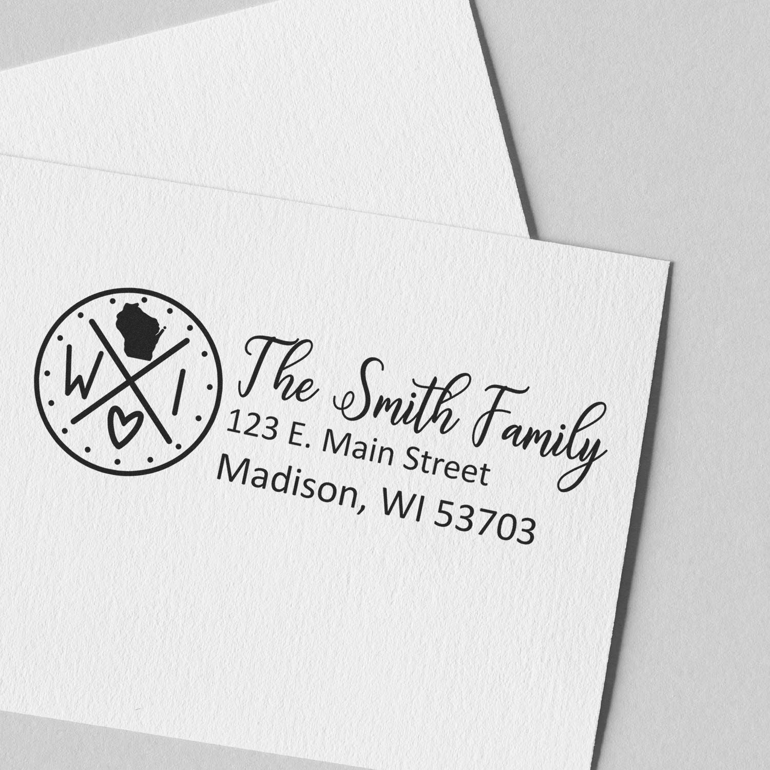 Slim Wisconsin Customized Pre-Inked Address Stamp on white paper, featuring a circular design with WI and a heart, alongside The Smith Family, 123 E. Main Street, Madison, WI 53703 in elegant font.