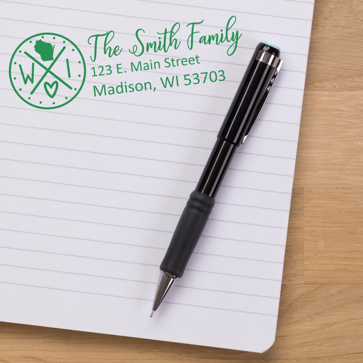 Wisconsin State Pride Customized Address Stamp on a notebook page with a pen. The stamp shows a family name and address in green, featuring a Wisconsin state outline and heart design.