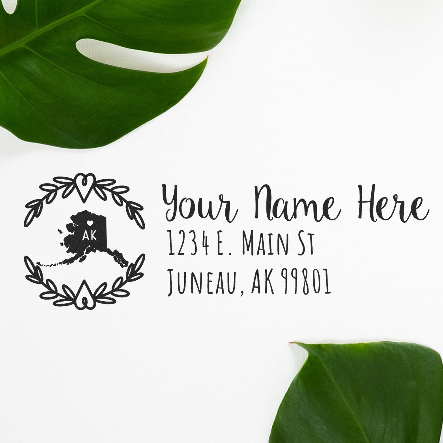 Alaska State Custom Return Address Stamp on white paper with a decorative Alaska map design, surrounded by green leaves. Text includes placeholder name and address in elegant font.