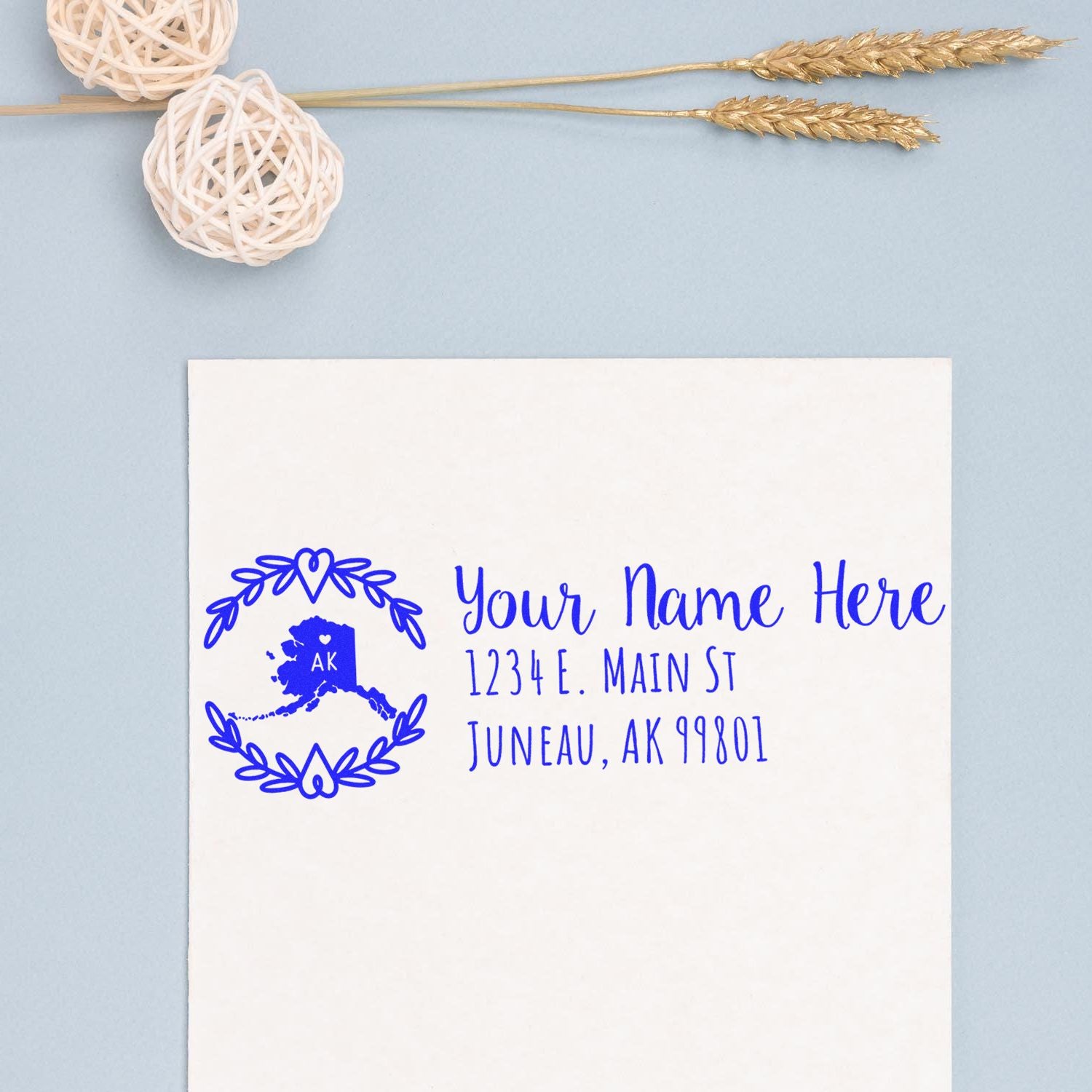 Alaska State Custom Return Address Stamp on white paper, featuring a blue Alaska map design with customizable text. Background includes decorative twine balls and wheat stalks on a light blue surface.