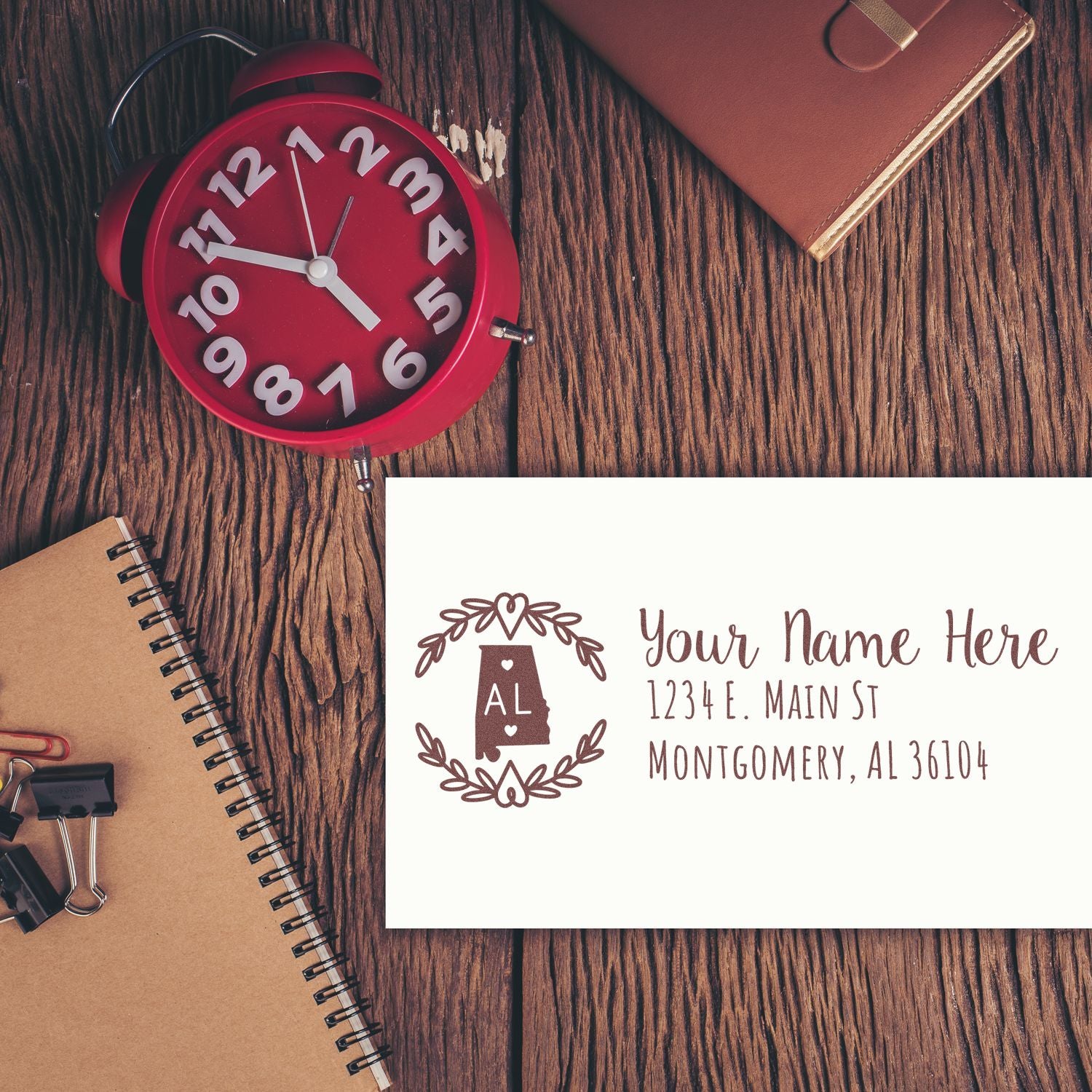 Alabama State Custom Return Address Stamp on an envelope, featuring a decorative state outline and address. Placed on a wooden desk with a red clock, notebook, and leather-bound journal.