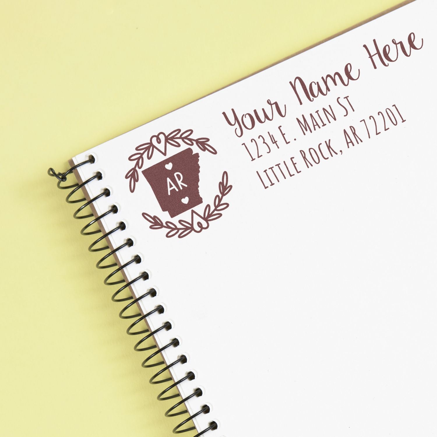 Arkansas State Custom Return Address Stamp on a spiral notebook, featuring a state outline with AR and decorative elements, alongside a sample address in elegant font.