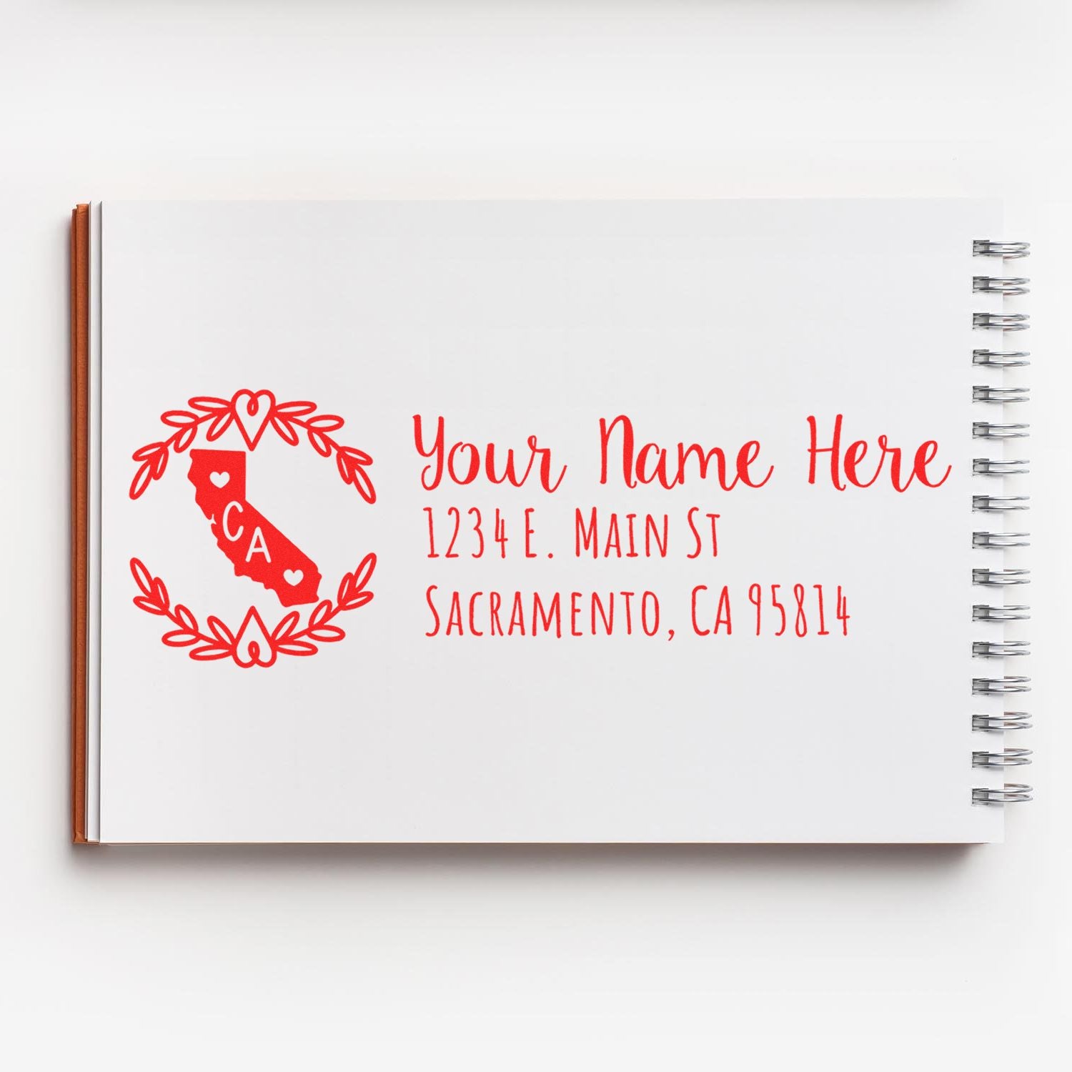 California State Custom Return Address Stamp in red ink on a white notebook, featuring a heart design with CA and sample address text.