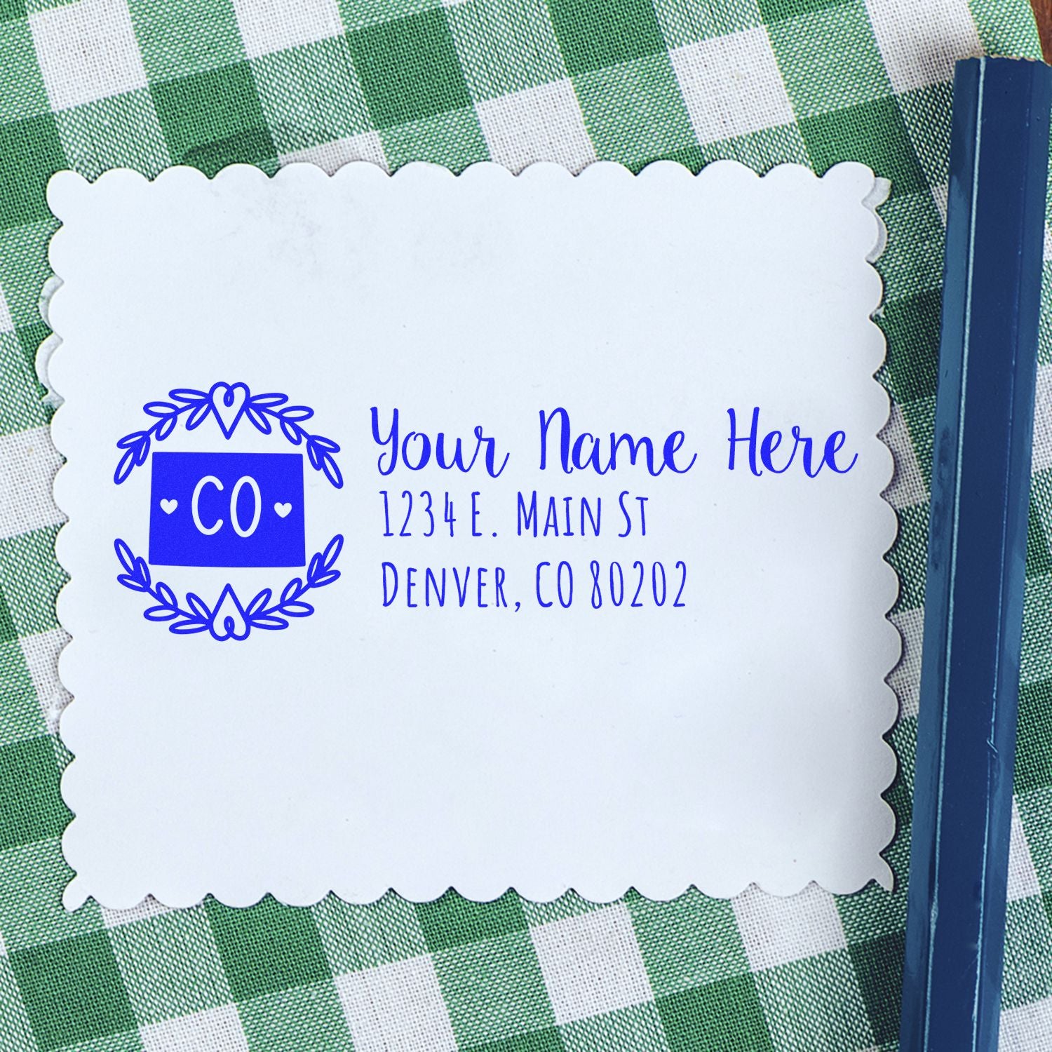 PSI Pre-Inked Colorado State Customized Address Stamp on a white card with scalloped edges, featuring a blue design and text, placed on a green and white checkered tablecloth.