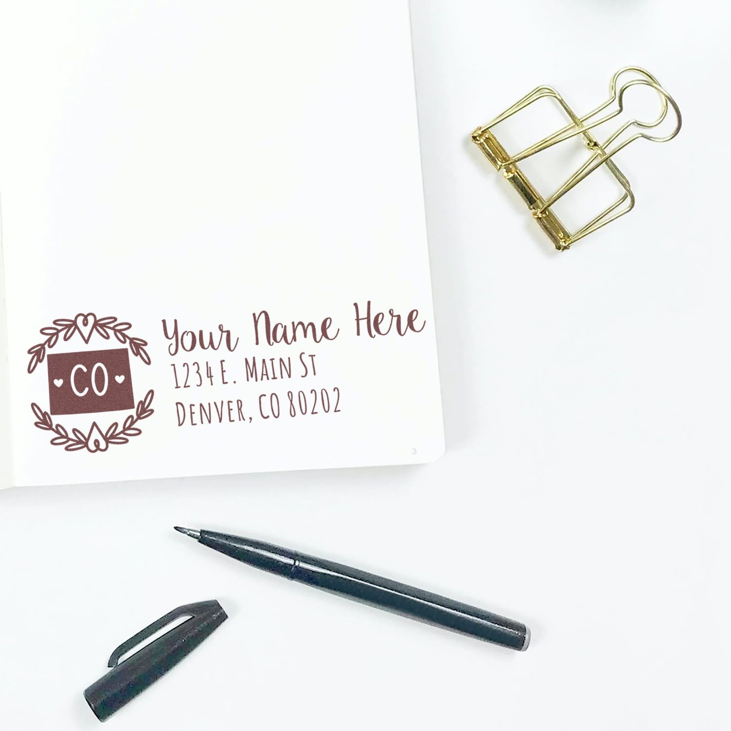 PSI Pre-Inked Colorado State Customized Address Stamp on white paper with a pen and gold binder clip nearby, featuring a decorative CO design and sample address text.