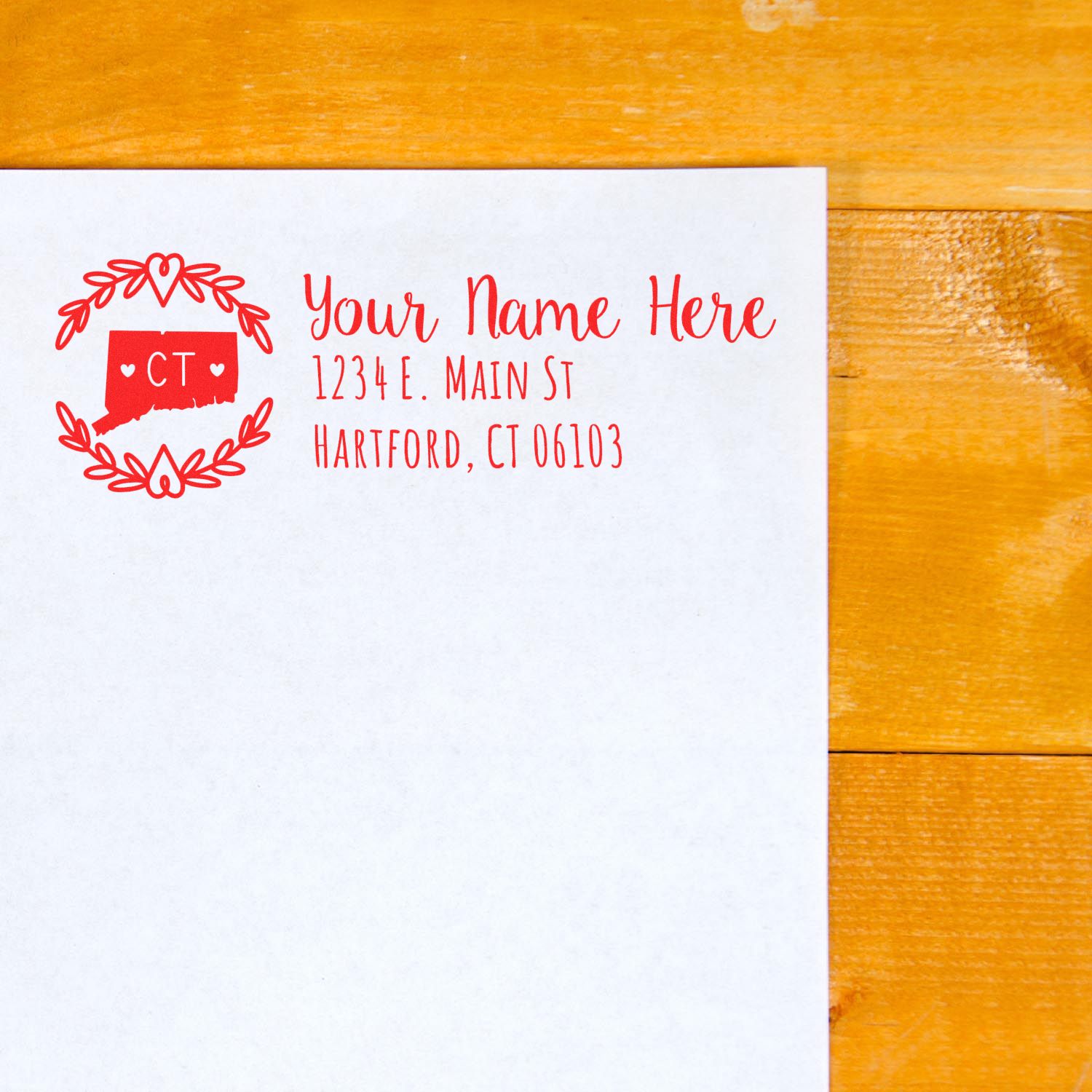 Self-Inking State Wreath of Connecticut Address Stamp in red ink on white paper, featuring a heart and wreath design with CT in the center, placed on a wooden surface.