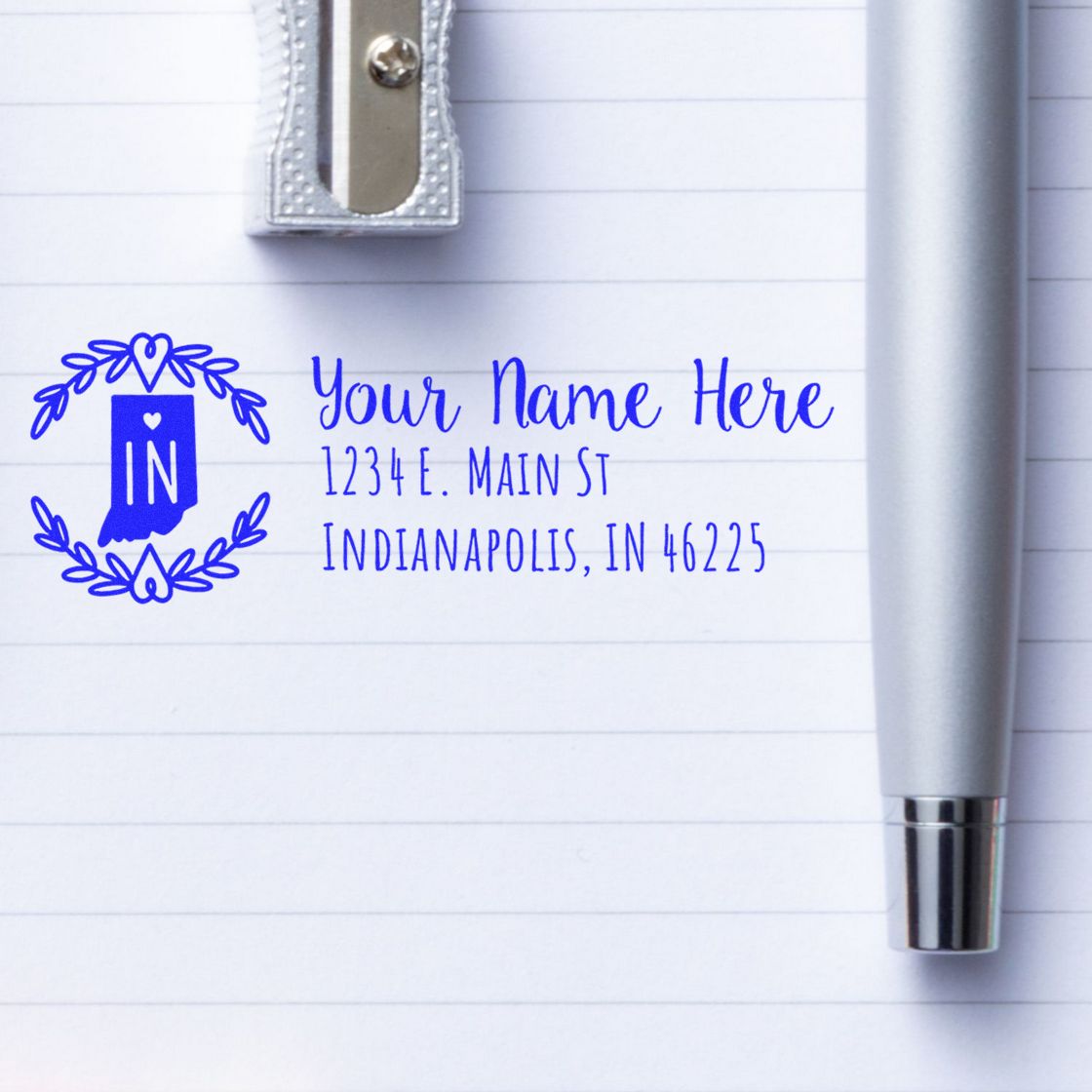 Indiana State Custom Return Address Stamp on lined paper with a pen and pencil sharpener. Features a blue state outline and personalized address text.