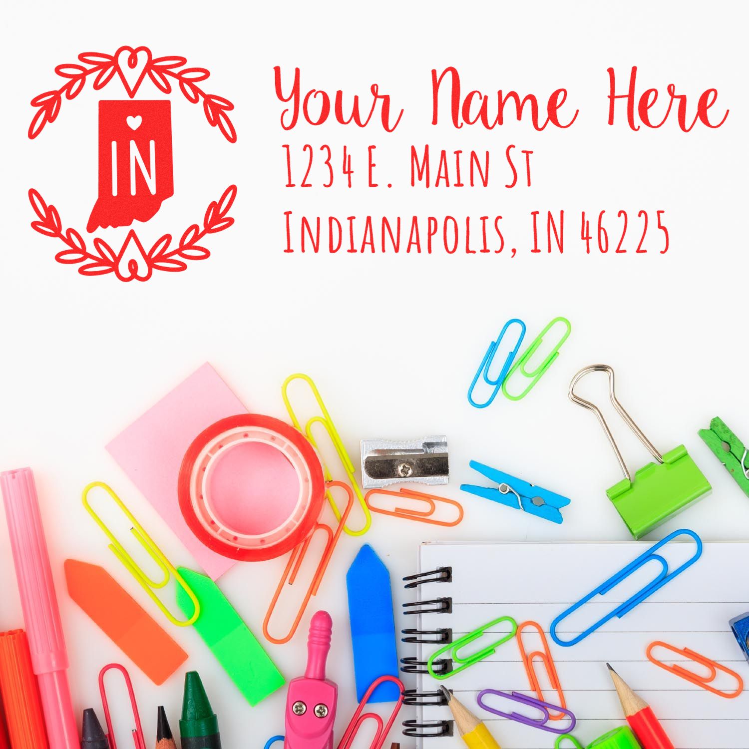 A colorful desk with stationery items surrounds a stamped envelope featuring the PSI Pre-Inked Indiana State Customized Address Stamp, displaying a sample address in red ink with a heart design.